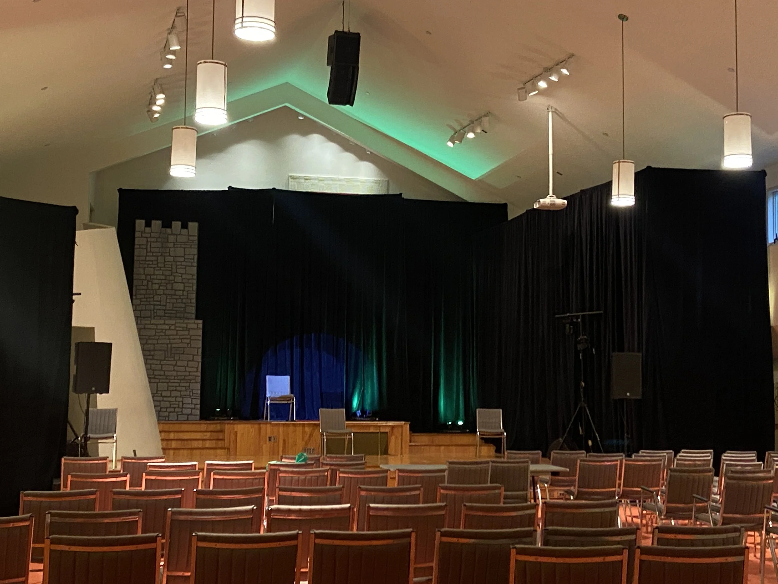 Empty theatre stage with black curtains, a single blue chair, and two chairs on the side, in front of an empty audience area with rows of wooden chairs. Stage lighting and a green-lit ceiling above.