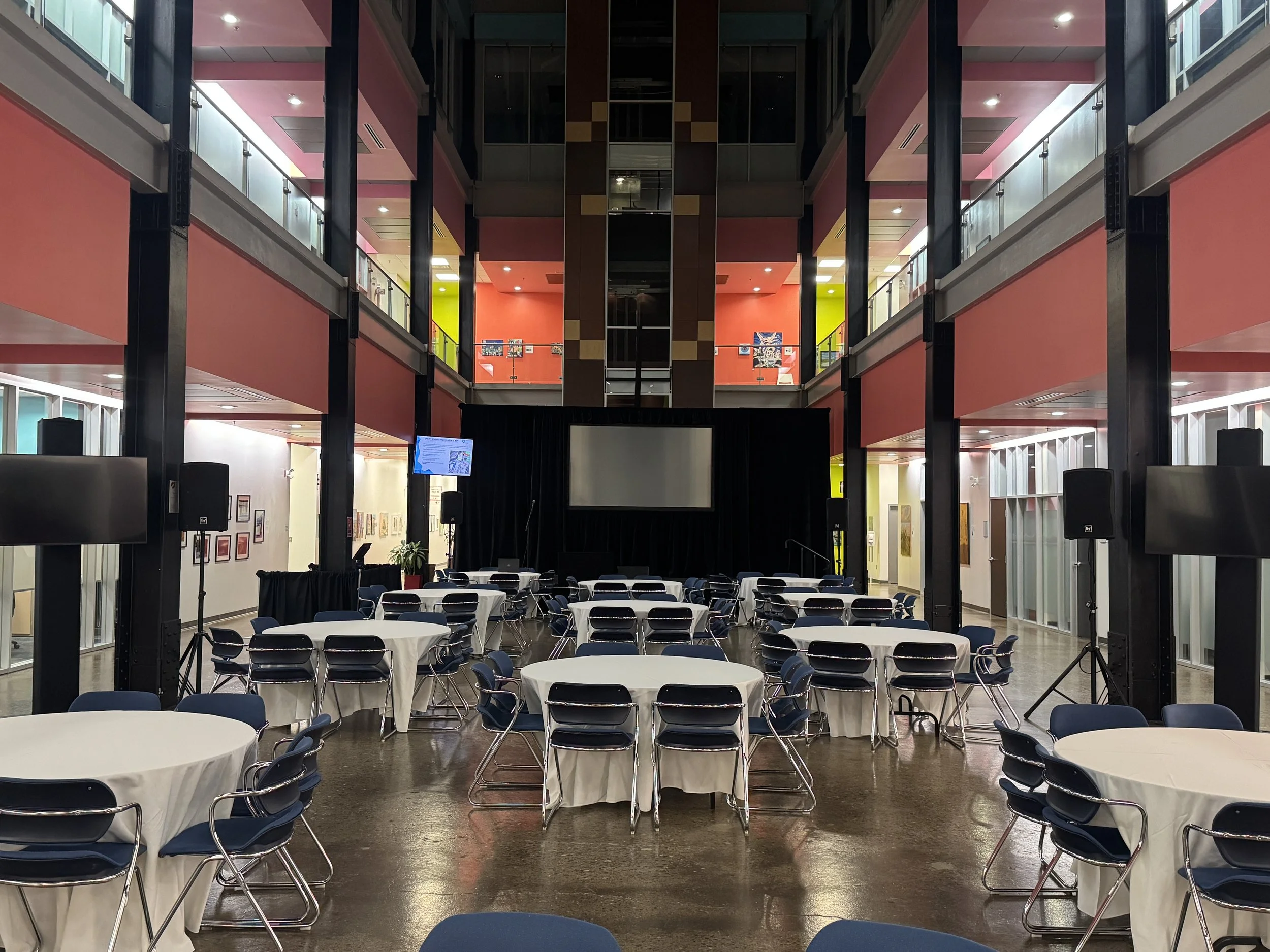 Event space with round tables covered in white tablecloths, stacked chairs, a large empty screen on stage, and audio equipment, in a modern multi-level building with colorful walls and artwork.