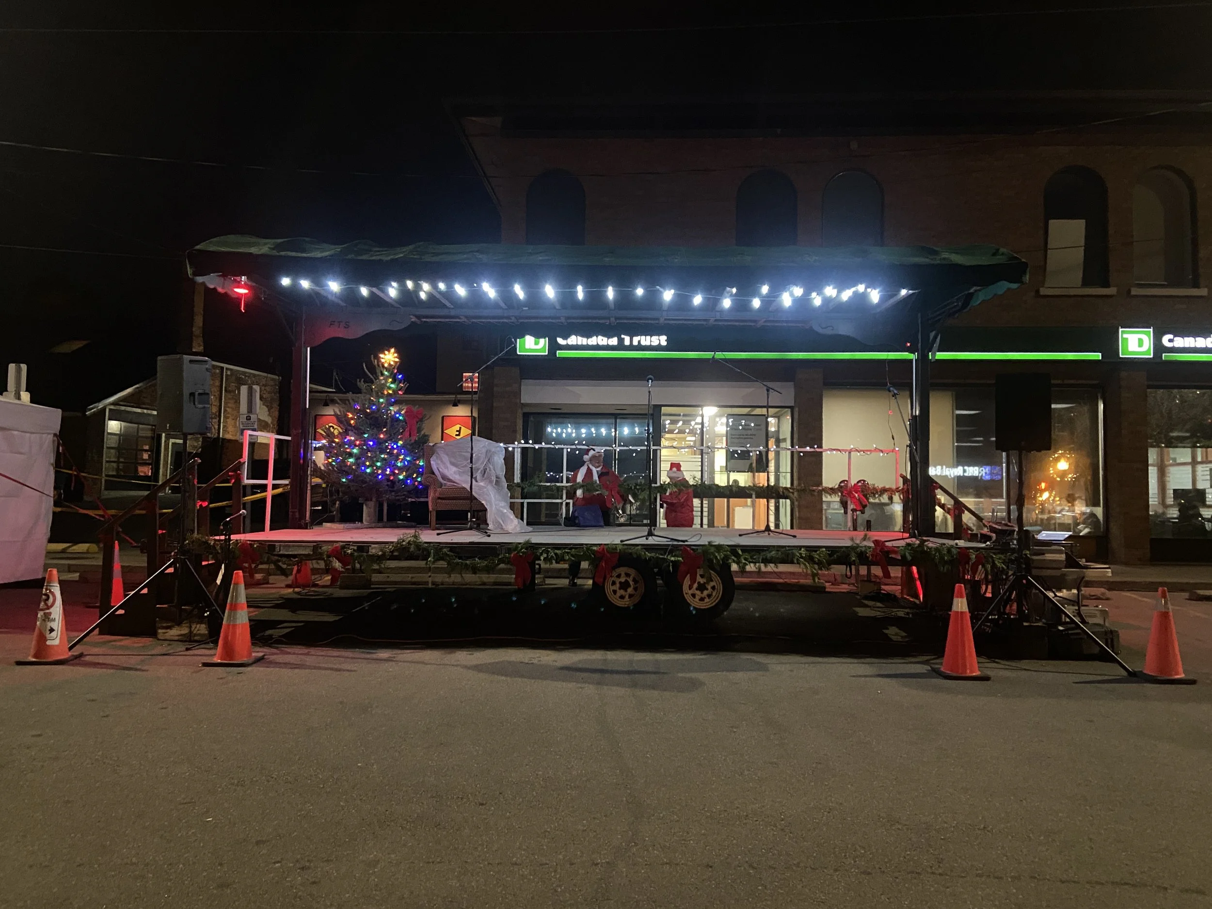 Outdoor Christmas decorated stage with a lit Christmas tree, Santa Claus figure, and holiday decorations, surrounded by traffic cones at night.