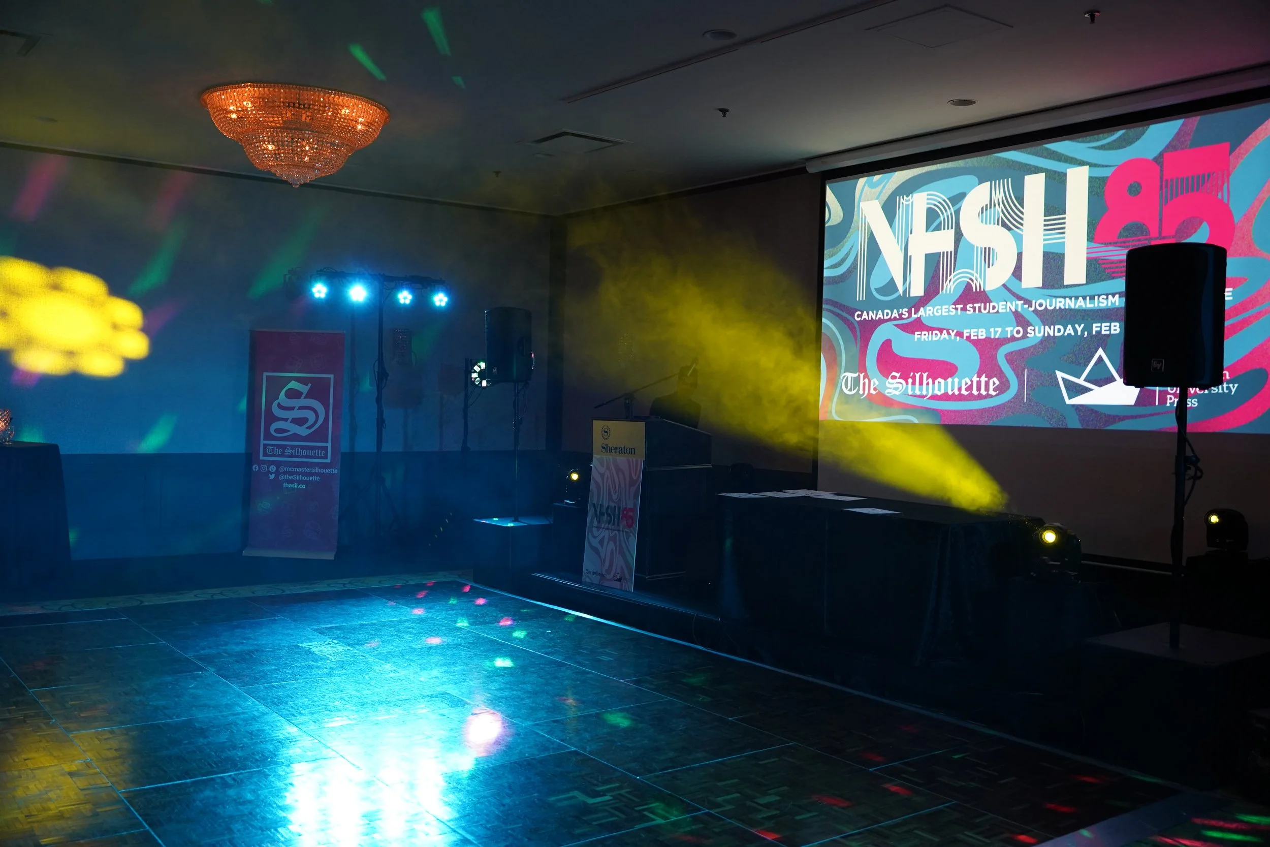 Event space with a large screen, blue and yellow lighting, a shiny dance floor, and a chandelier hanging from the ceiling.