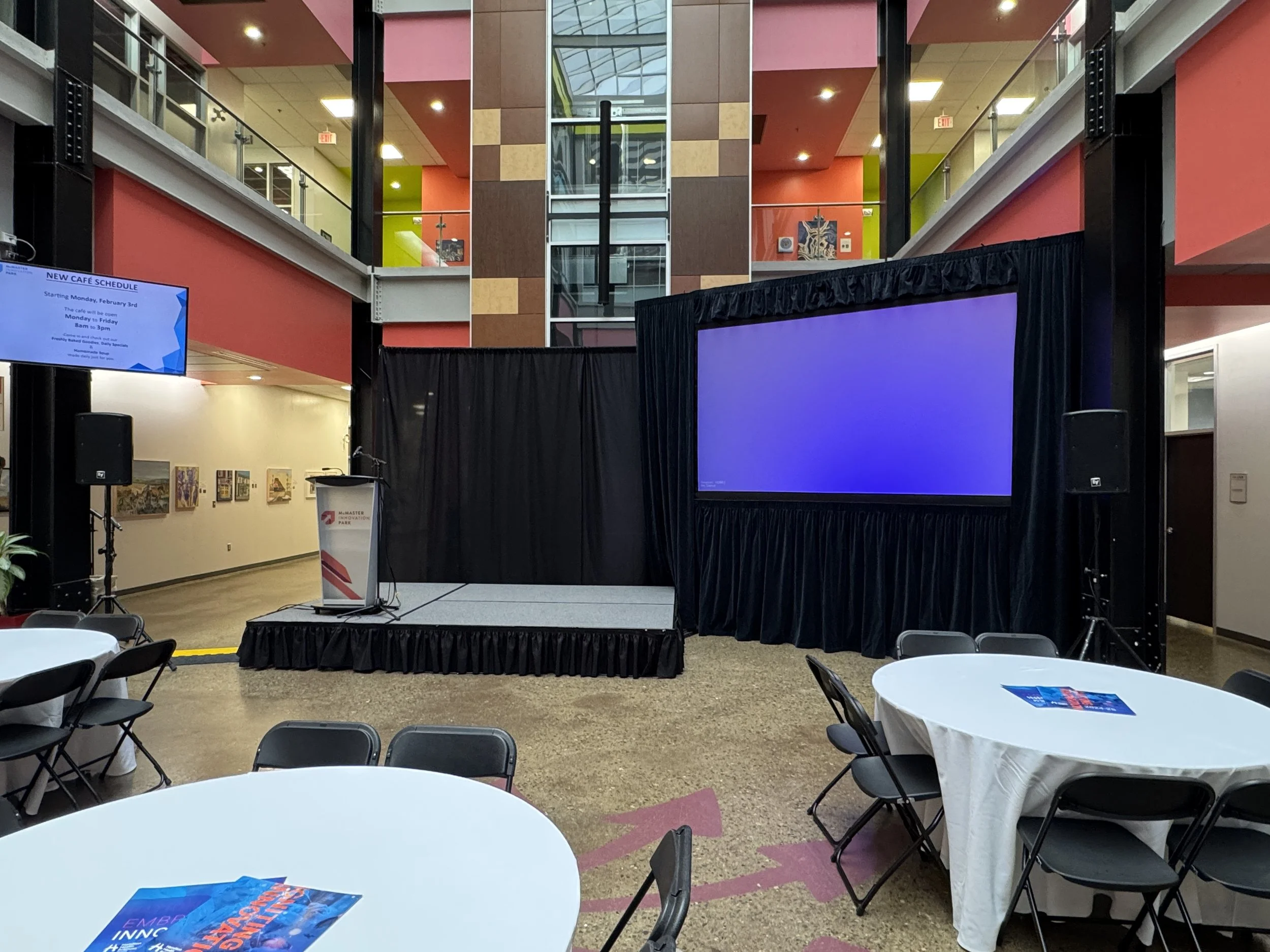Event space with round tables covered with white tablecloths, black chairs, a small stage with a podium, large screen on the right, black curtains, and a second screen on the left displaying a schedule, inside a multi-level building.