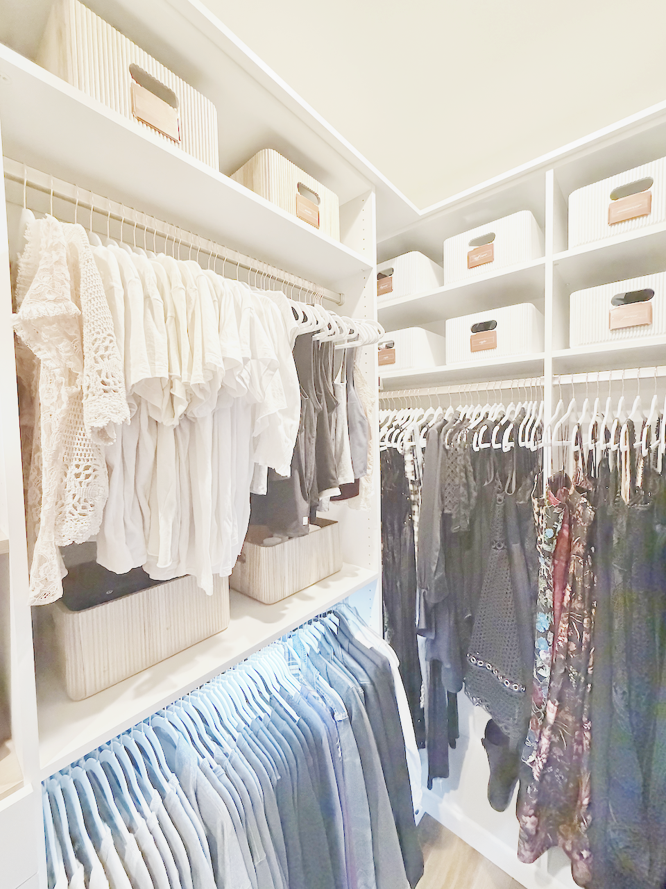 An organized client closet with bright lighting, white shelves, color coordinated clothes on white hangers with white bins on the shelves above the hanging clothes.