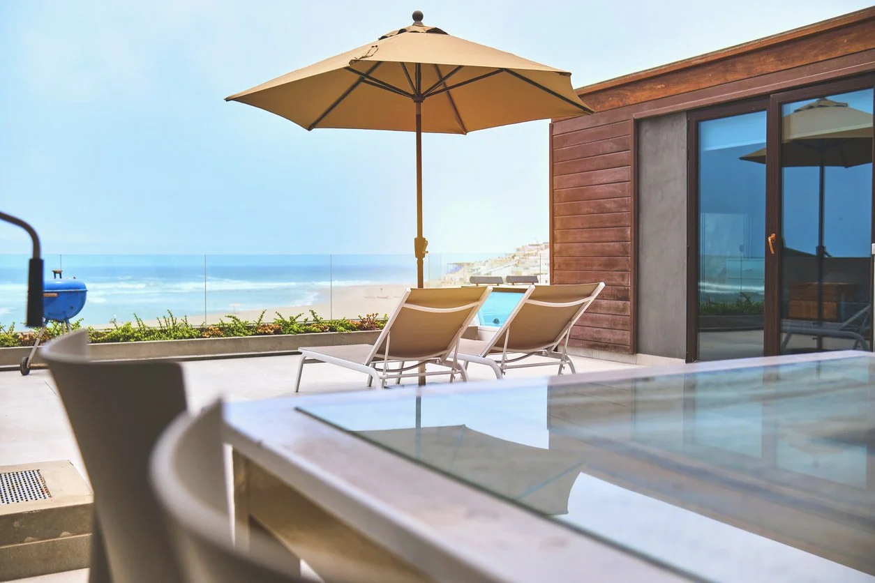 Ocean-view vacation home patio with lounge chairs and umbrella overlooking the beach, prepared and ready for arrival.