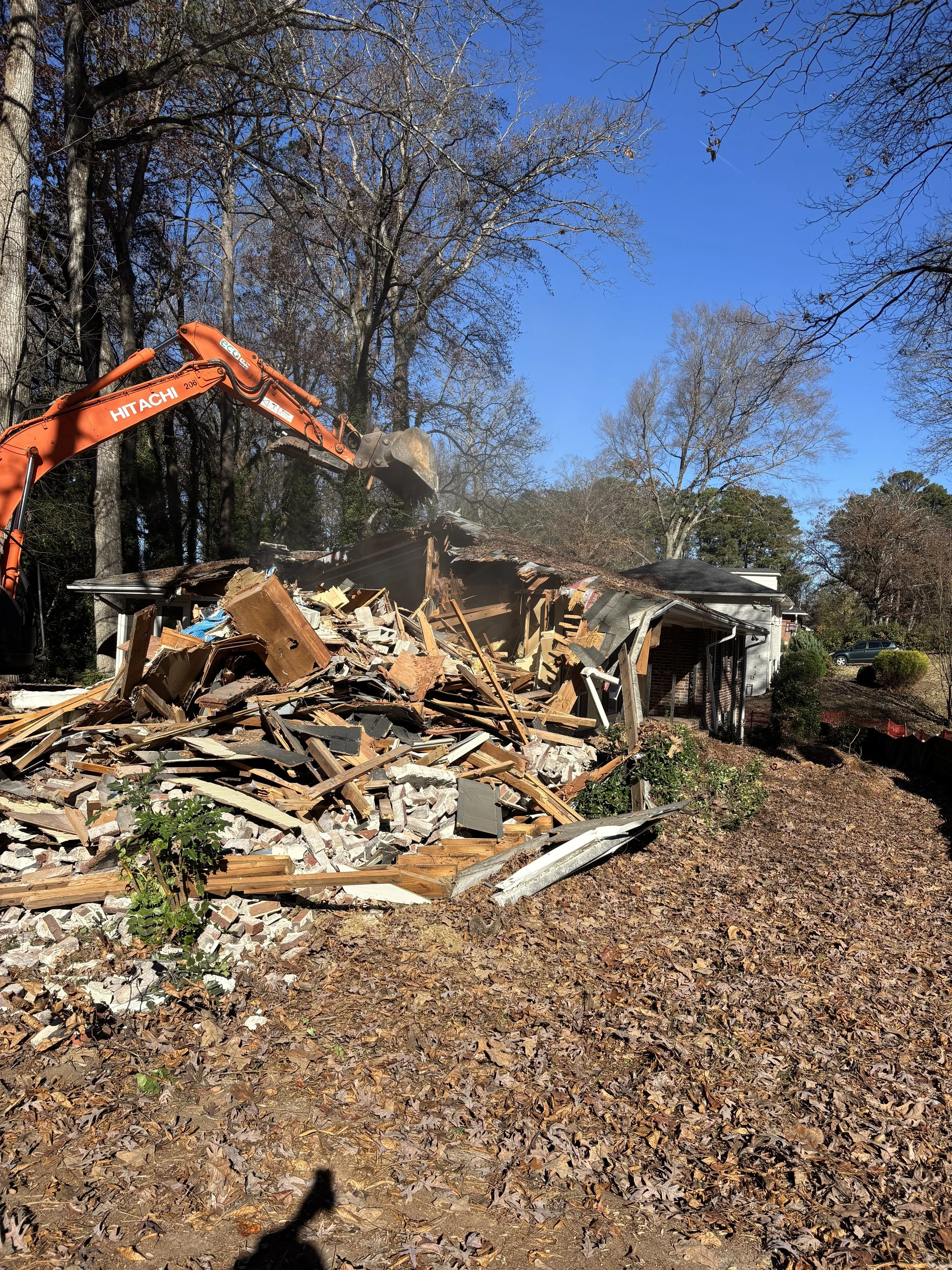 A house is being demolished with an orange excavator, with debris and wood piled up in front, on a cloudy day with clear blue sky and fall leaves on the ground.