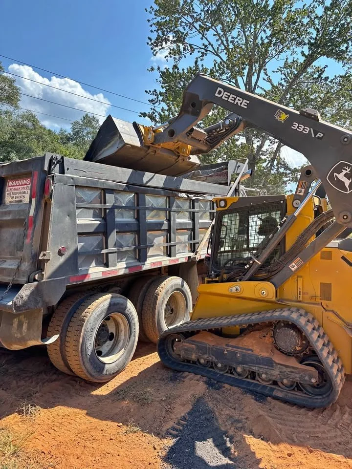 Construction site with a John Deere compact excavator loading dirt into a dump truck under a blue sky with trees.