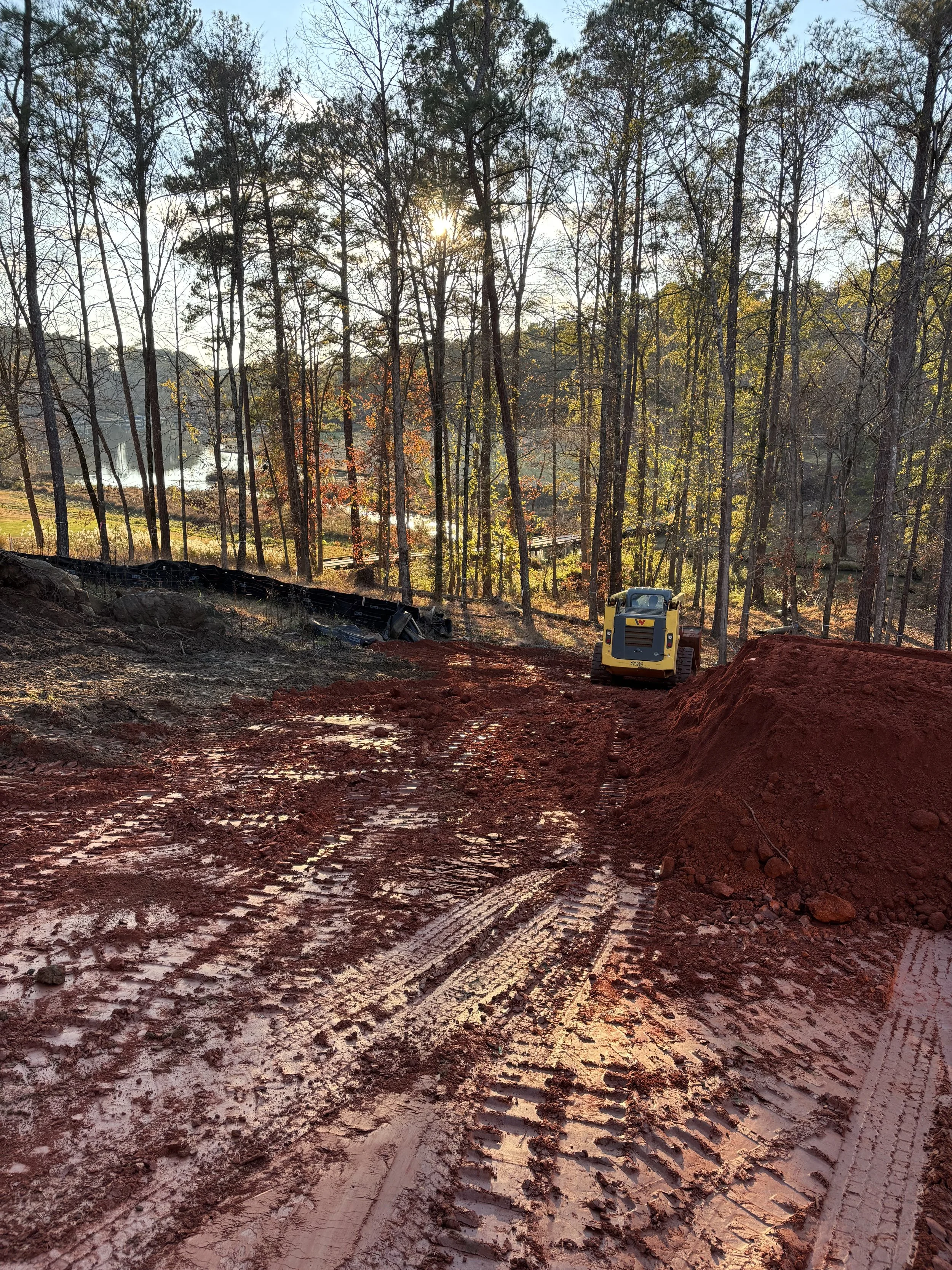 Construction site with muddy red dirt, tire tracks, and a small yellow bulldozer surrounded by trees and a lake in the background.