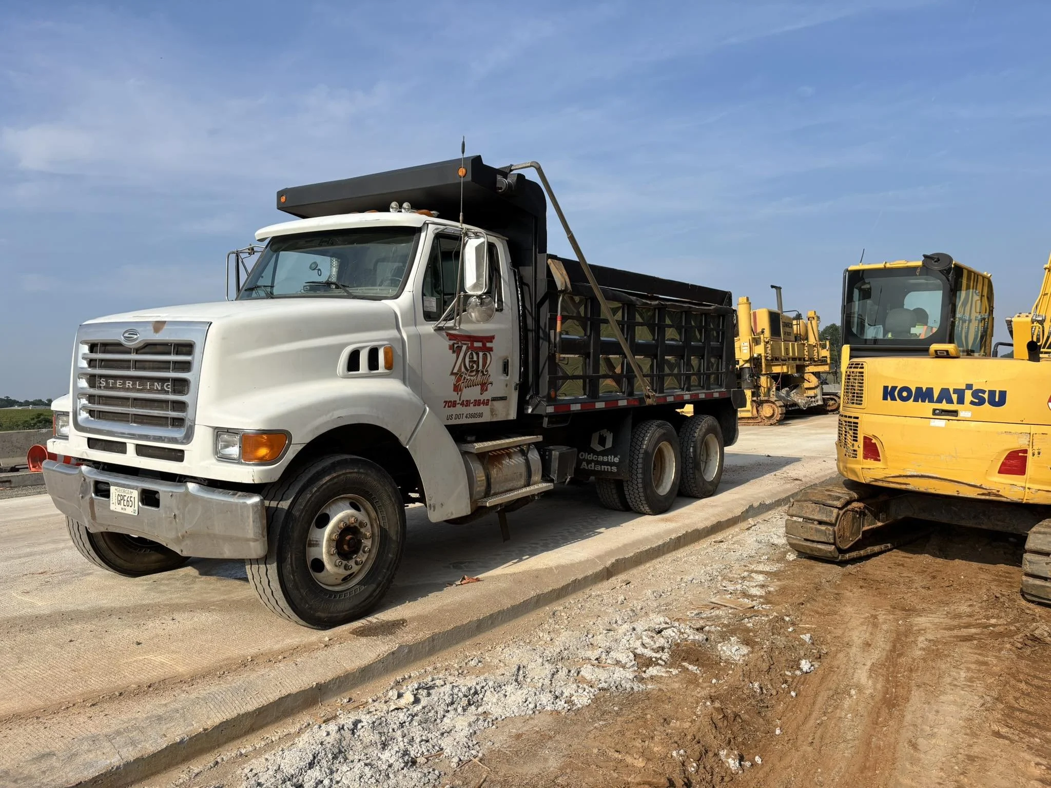 A construction site with a white dump truck and a yellow Komatsu excavator parked on dirt, against a clear blue sky.