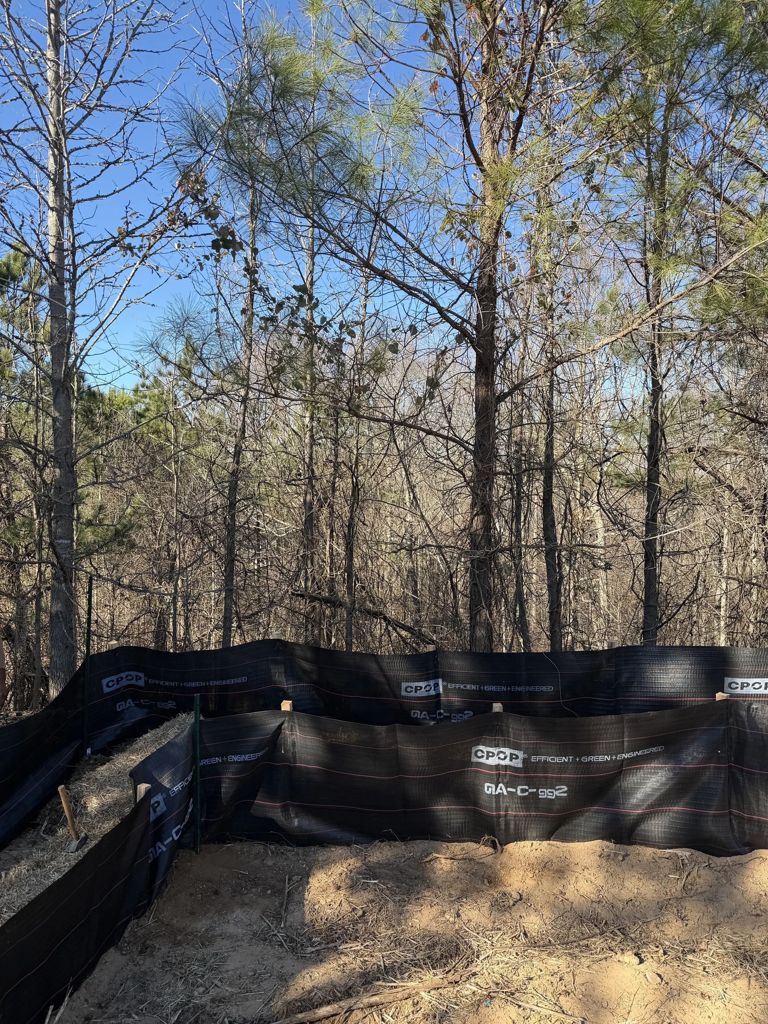 A construction site with a black silt fence, wooden stakes, and soil in a wooded area with leafless and evergreen trees under a clear blue sky.