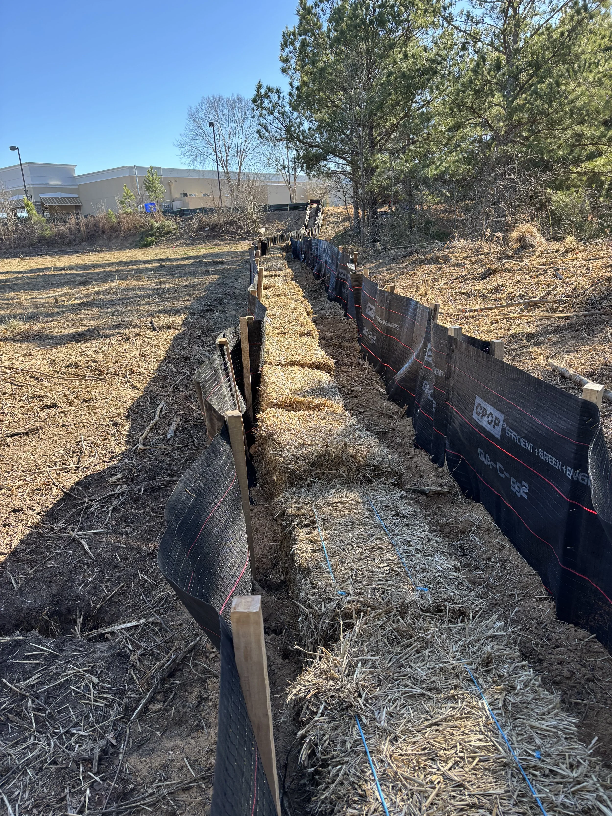 A landscaped outdoor area with a trench filled with straw bales, bordered by black silt fencing and wooden stakes, under a clear blue sky.
