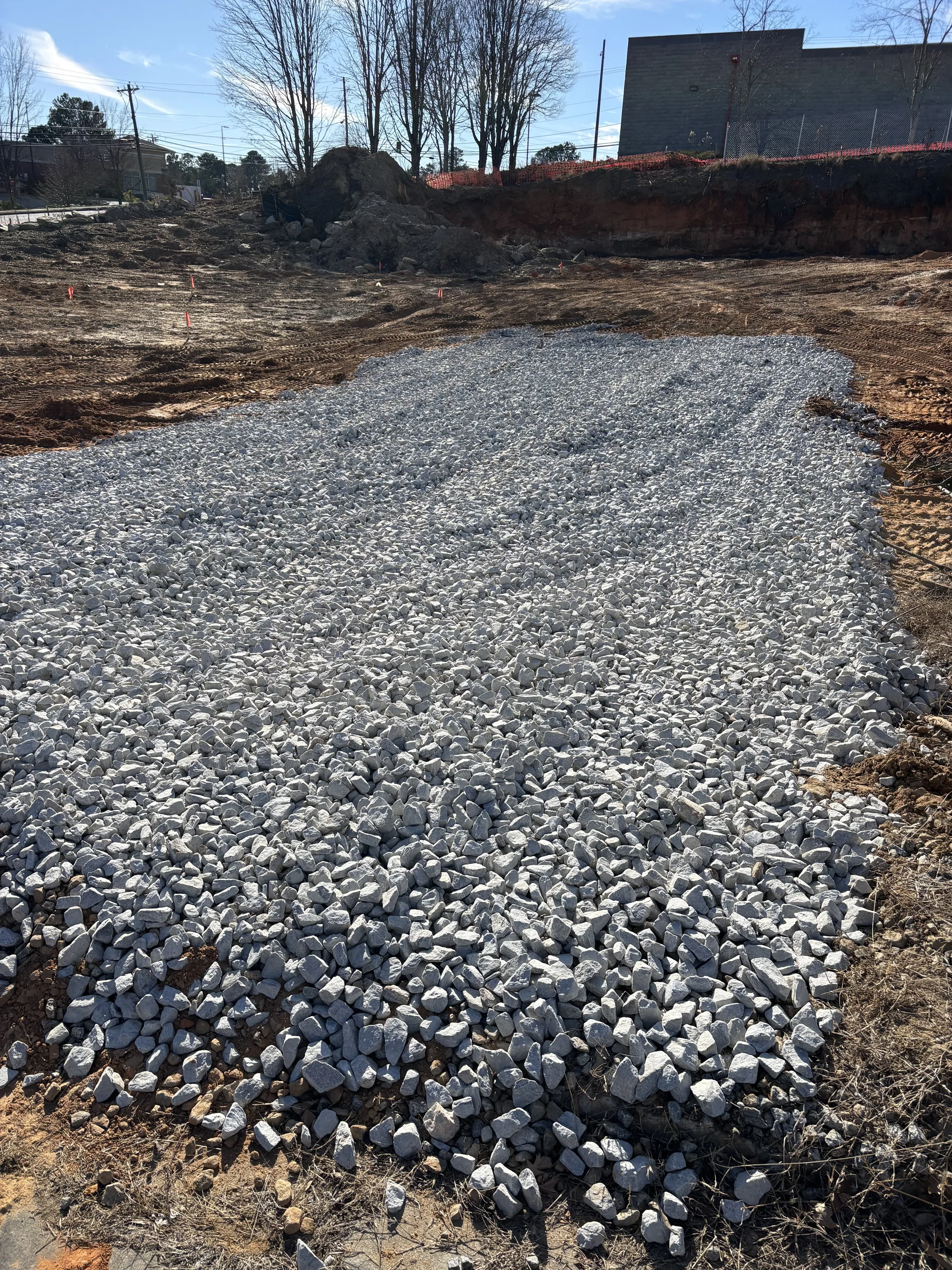 Construction site with a bed of gravel on the ground, partly excavated earth, and a building in the background with trees and power lines.