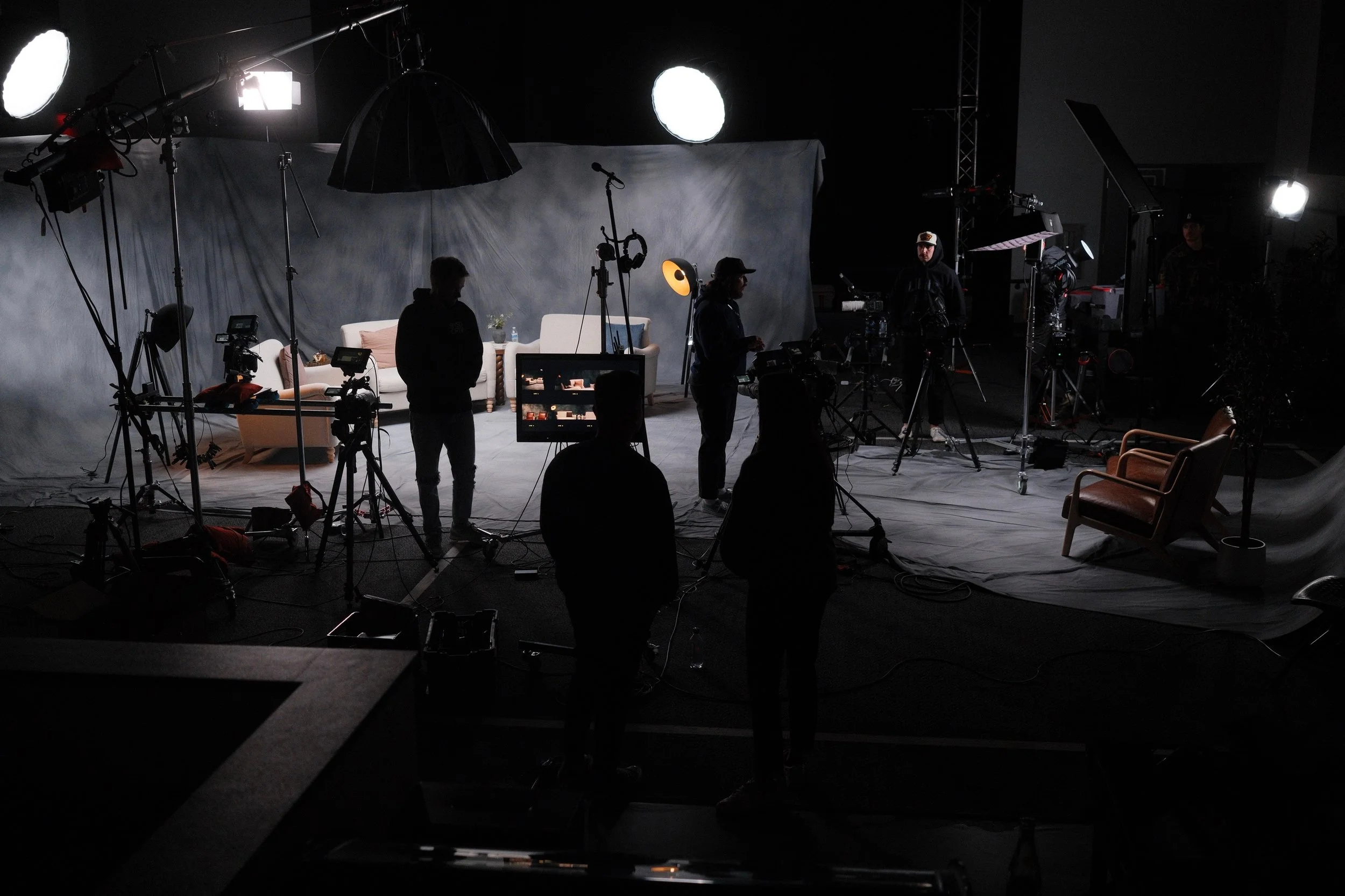 Behind the scenes of a video shoot with professional lighting, cameras, and crew preparing the set with a sofa, armchair, and backdrop.