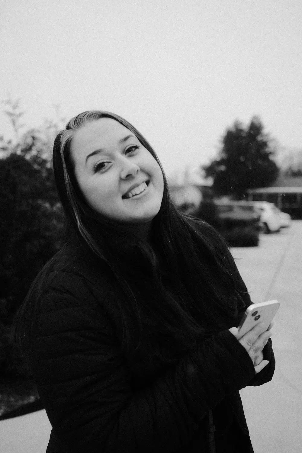 A woman with long dark hair smiling and holding a smartphone outdoors, with cars and trees in the background, in black and white.