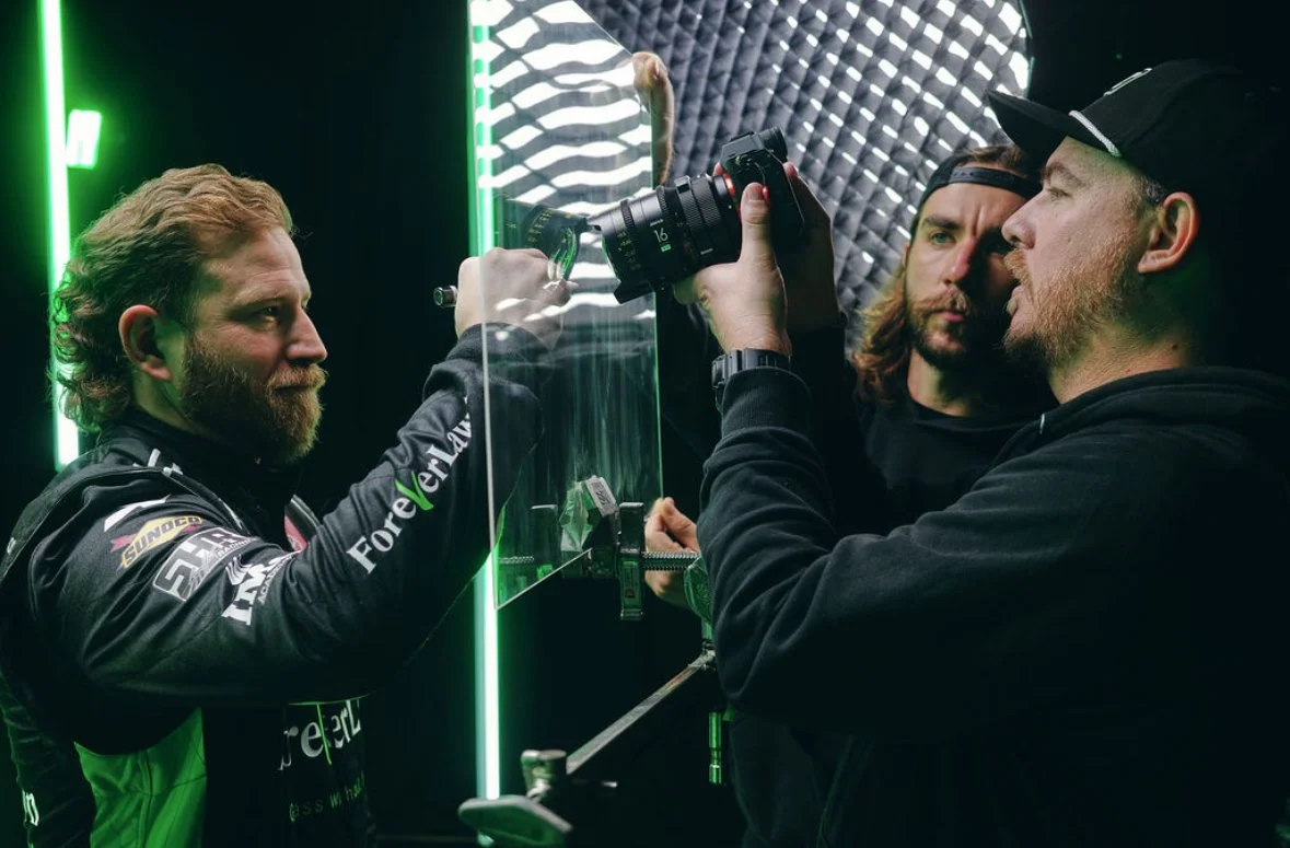 Three men working together in a studio with soundproofing and neon lights. One man in racing gear is holding a clear glass panel, while the other two men, wearing black hoodies and caps, are filming or taking photos of him with a professional camera.