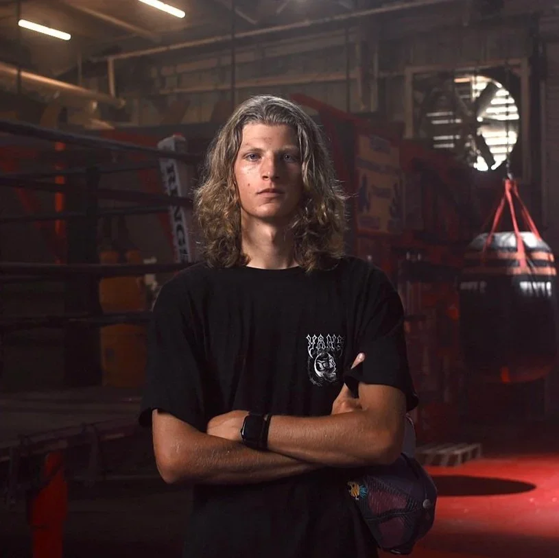 Young man with long hair crossing arms in a boxing gym with boxing equipment and punching bags in the background.
