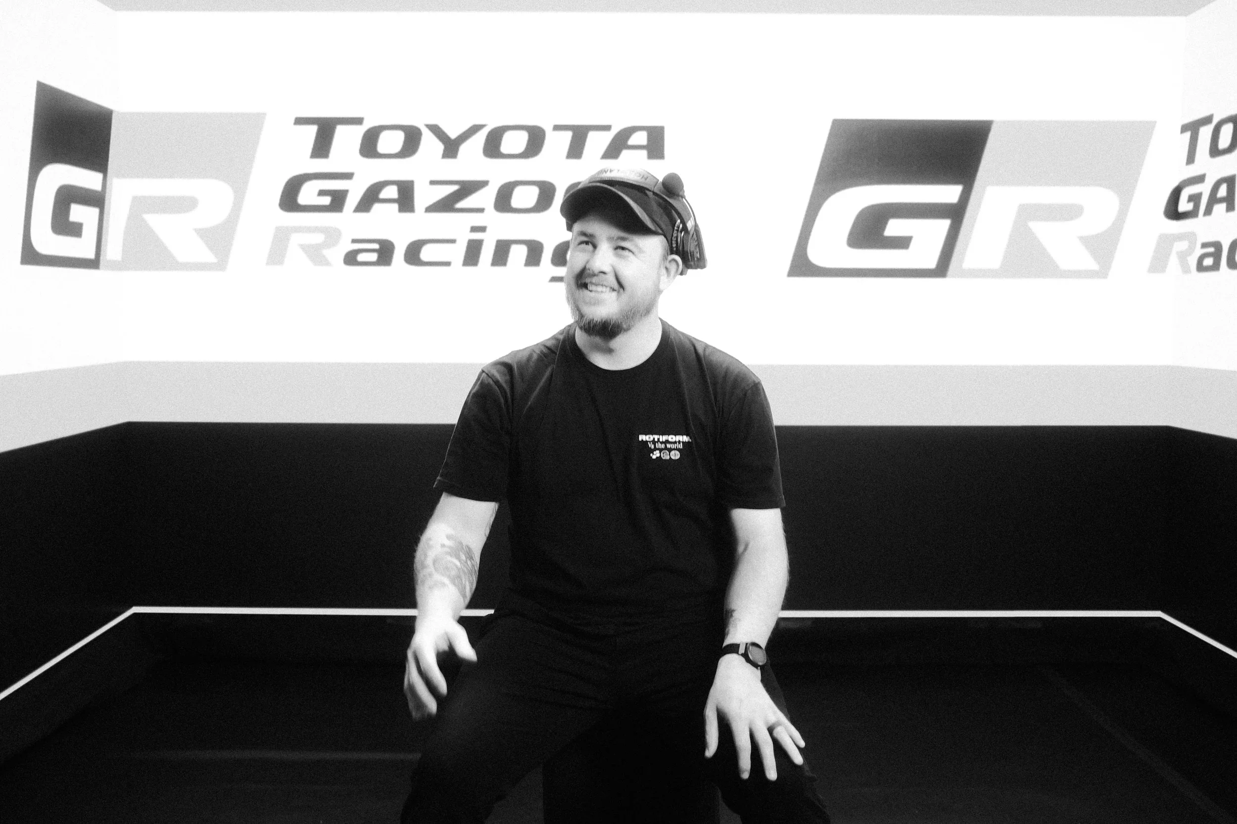 A man with a beard and a cap, wearing a black T-shirt, is sitting on a platform at a racing event, with a backdrop displaying the logos of Toyota Gazoo Racing and GR.