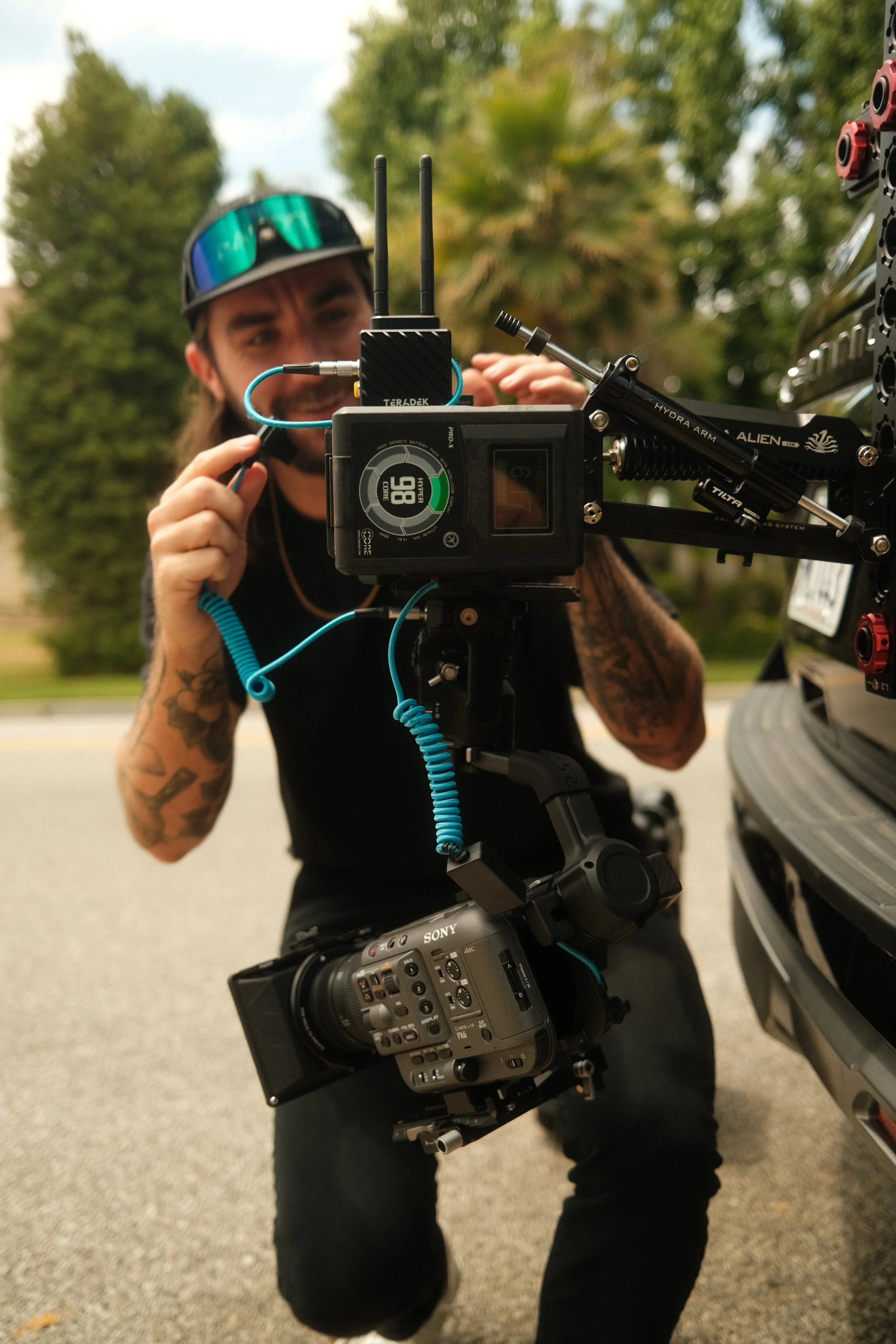 A man with tattoos and wearing sunglasses is operating a professional video camera mounted on a stabilizer near a vehicle outdoors with trees in the background.