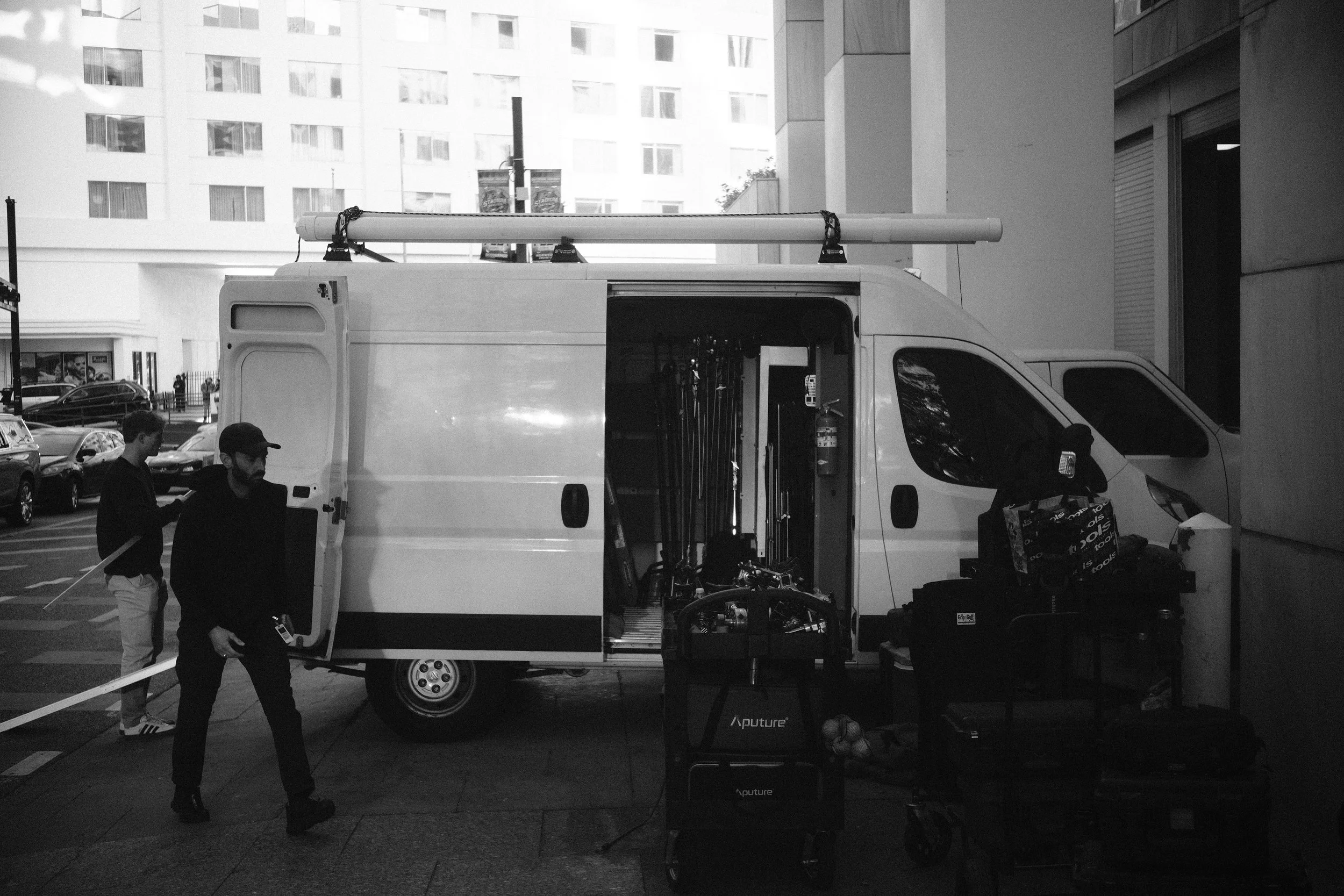 A service van parked on a city street with equipment inside and outside, including carts and tools, while two men stand nearby.
