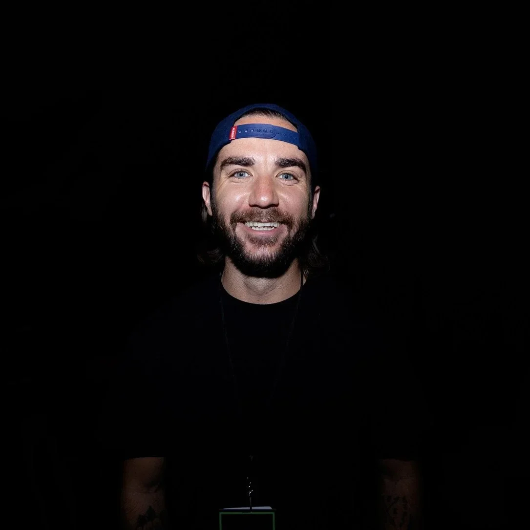 A smiling man with a beard, wearing a dark cap backwards, black shirt, and a lanyard, against a black background.