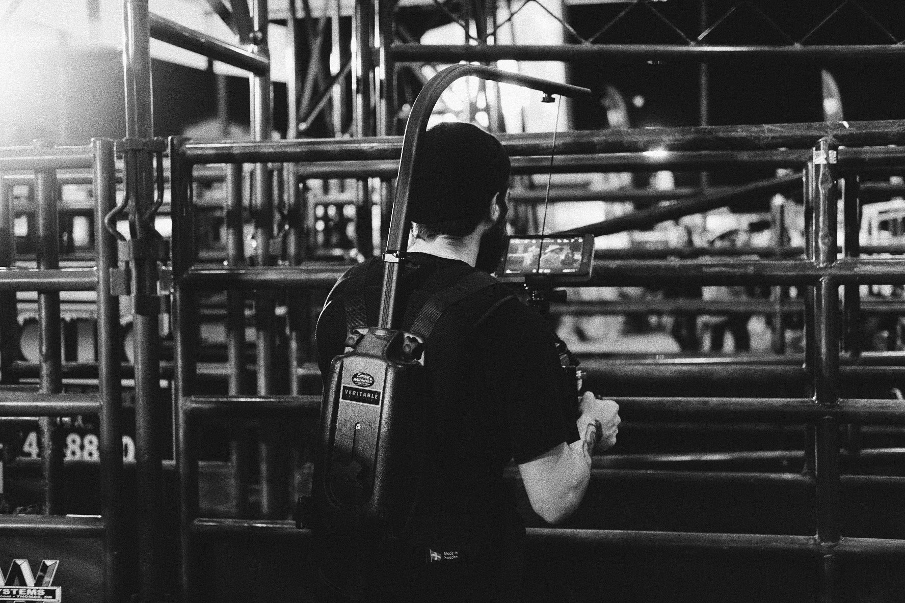 A person operating a camera mounted on a shoulder rig, filming or photographing in an indoor setting with metal shelves or scaffolding around them, in black and white.