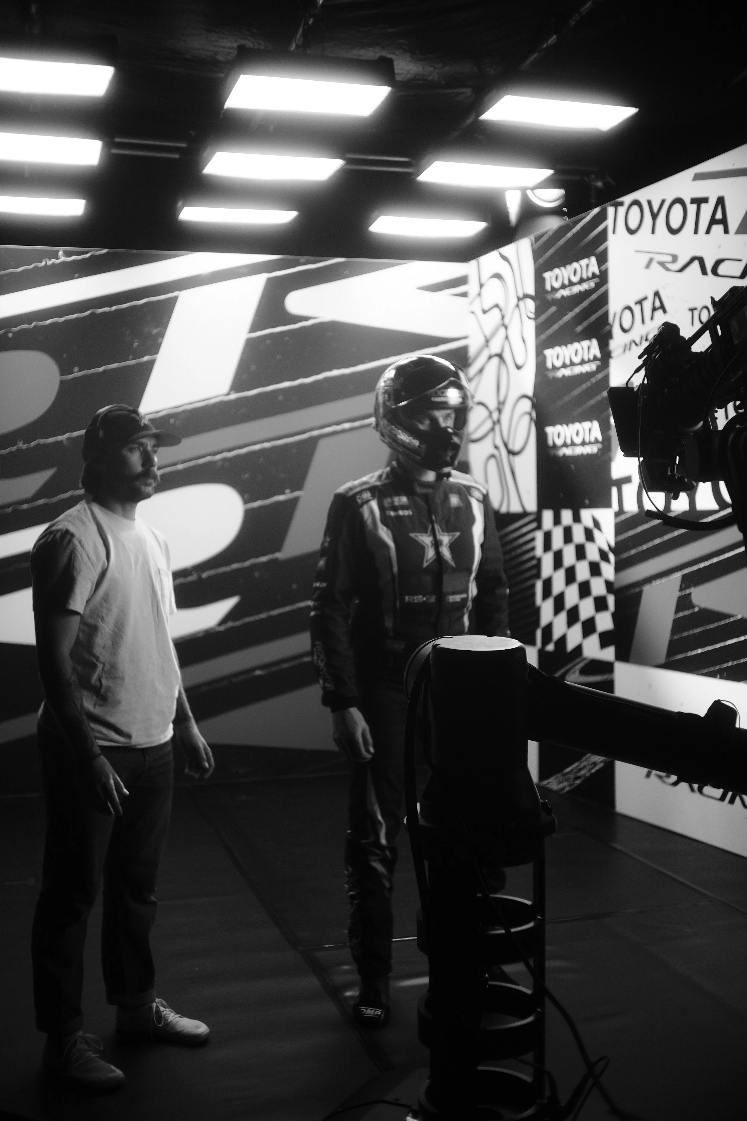 Black and white image of a man in racing suit and helmet standing next to a crew member in casual clothing, with a backdrop displaying sponsors and racing graphics. They are in a studio or stage setting with bright ceiling lights.
