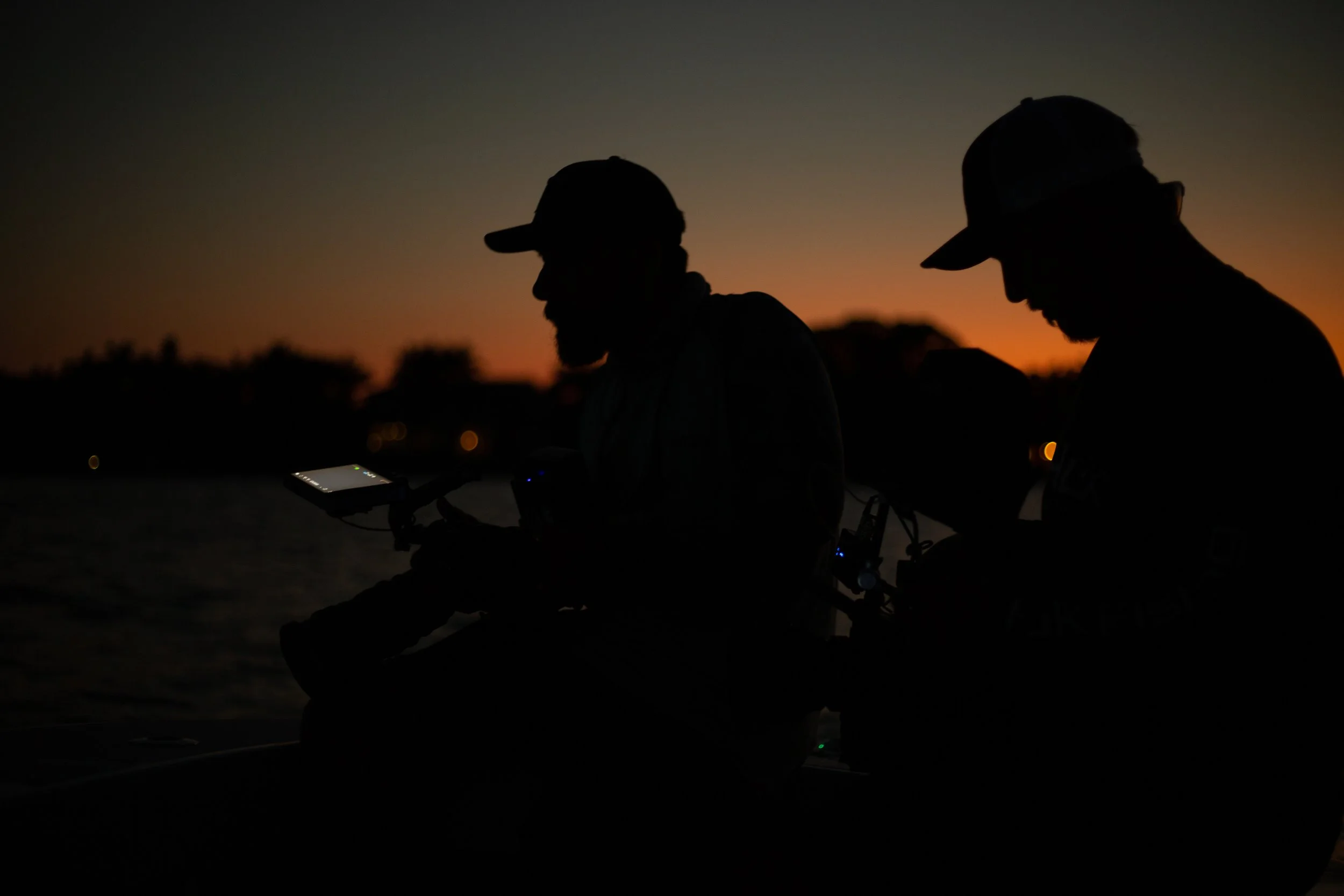 Two silhouetted men wearing hats and glasses sitting outdoors at sunset, looking at a device.