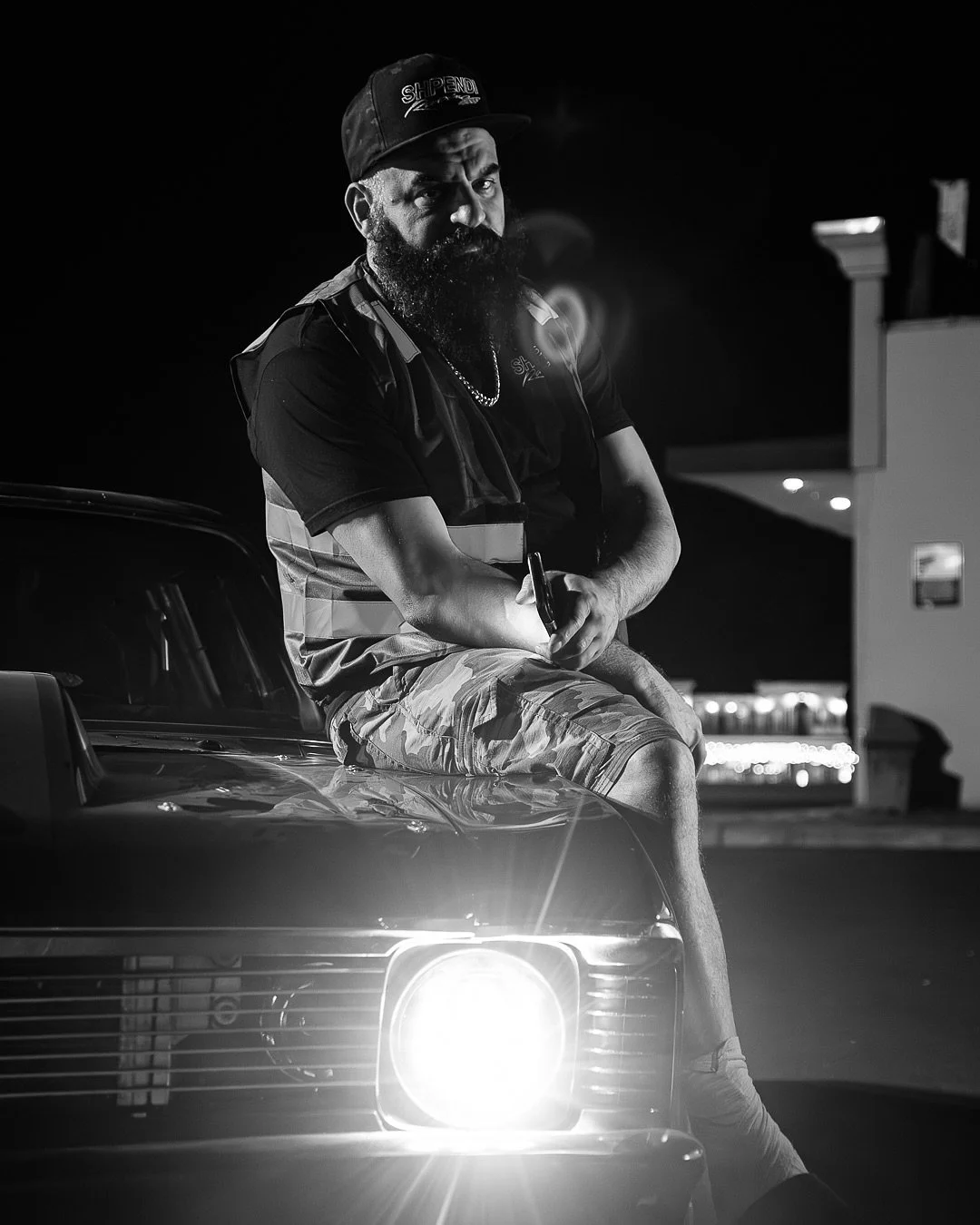 A man with a beard wearing a cap and casual clothing sitting on the hood of a car at night.