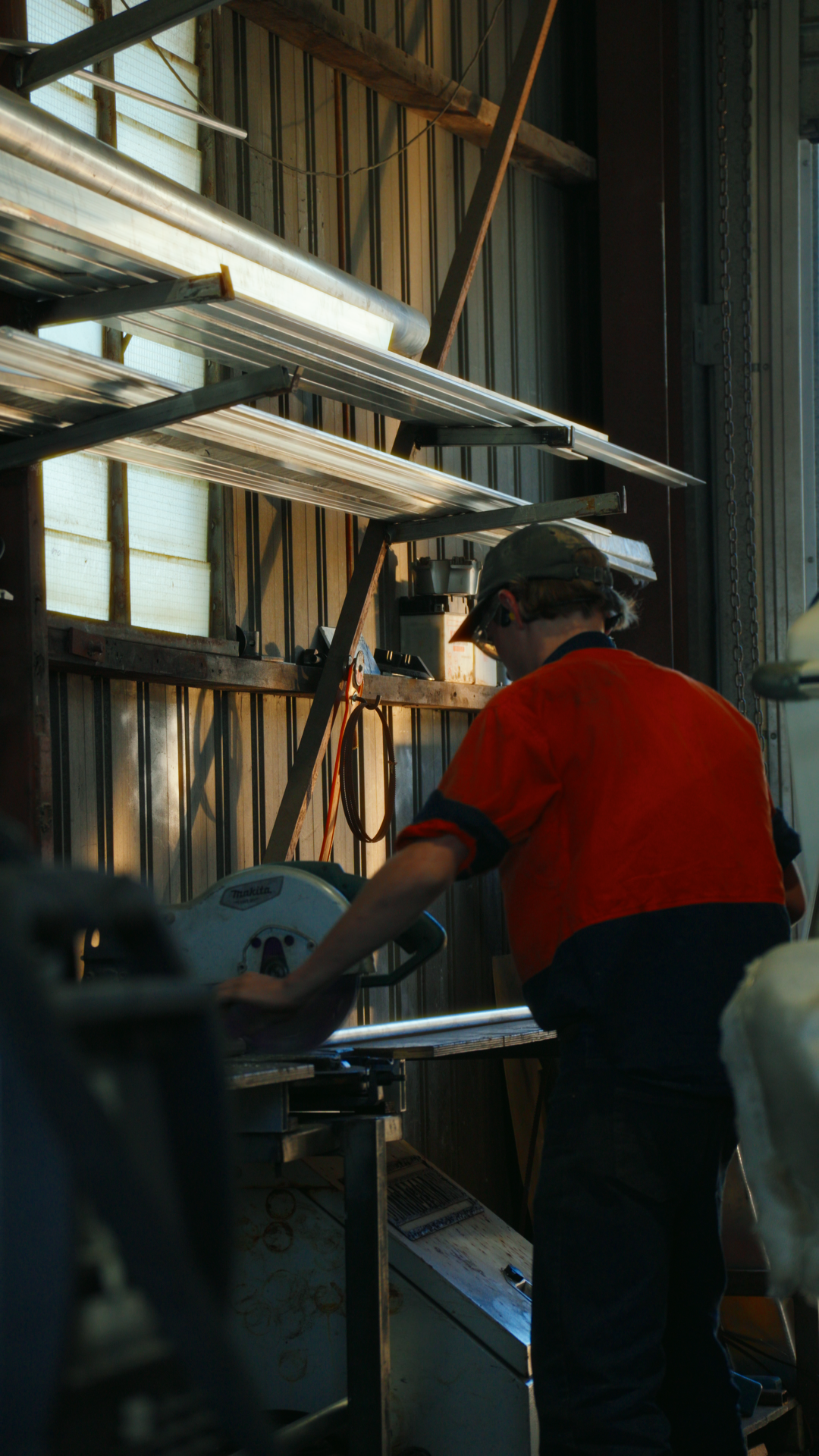 A person wearing an orange and black work uniform and a cap is using a saw in a workshop with metal shelves and tools.