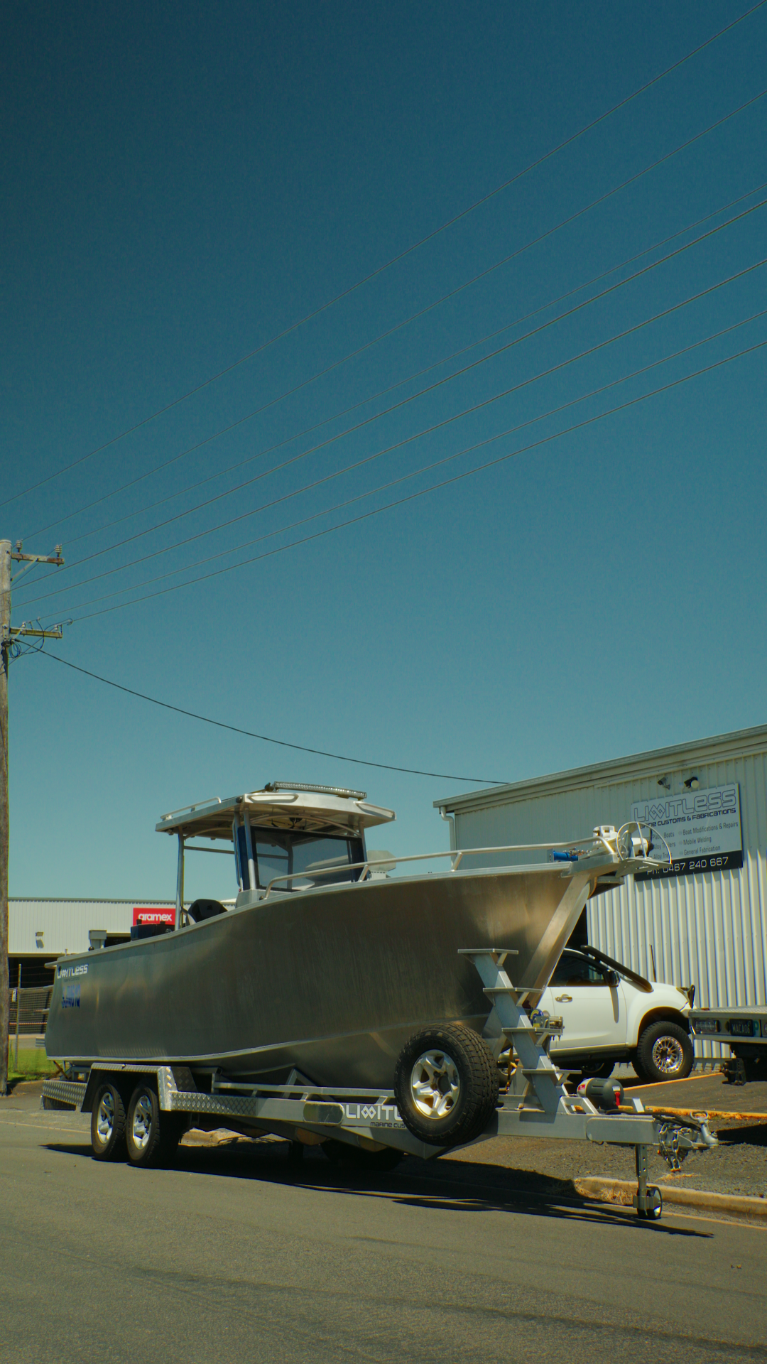 A boat on a trailer parked in an industrial lot with a white vehicle and metal building in the background, under a clear blue sky.