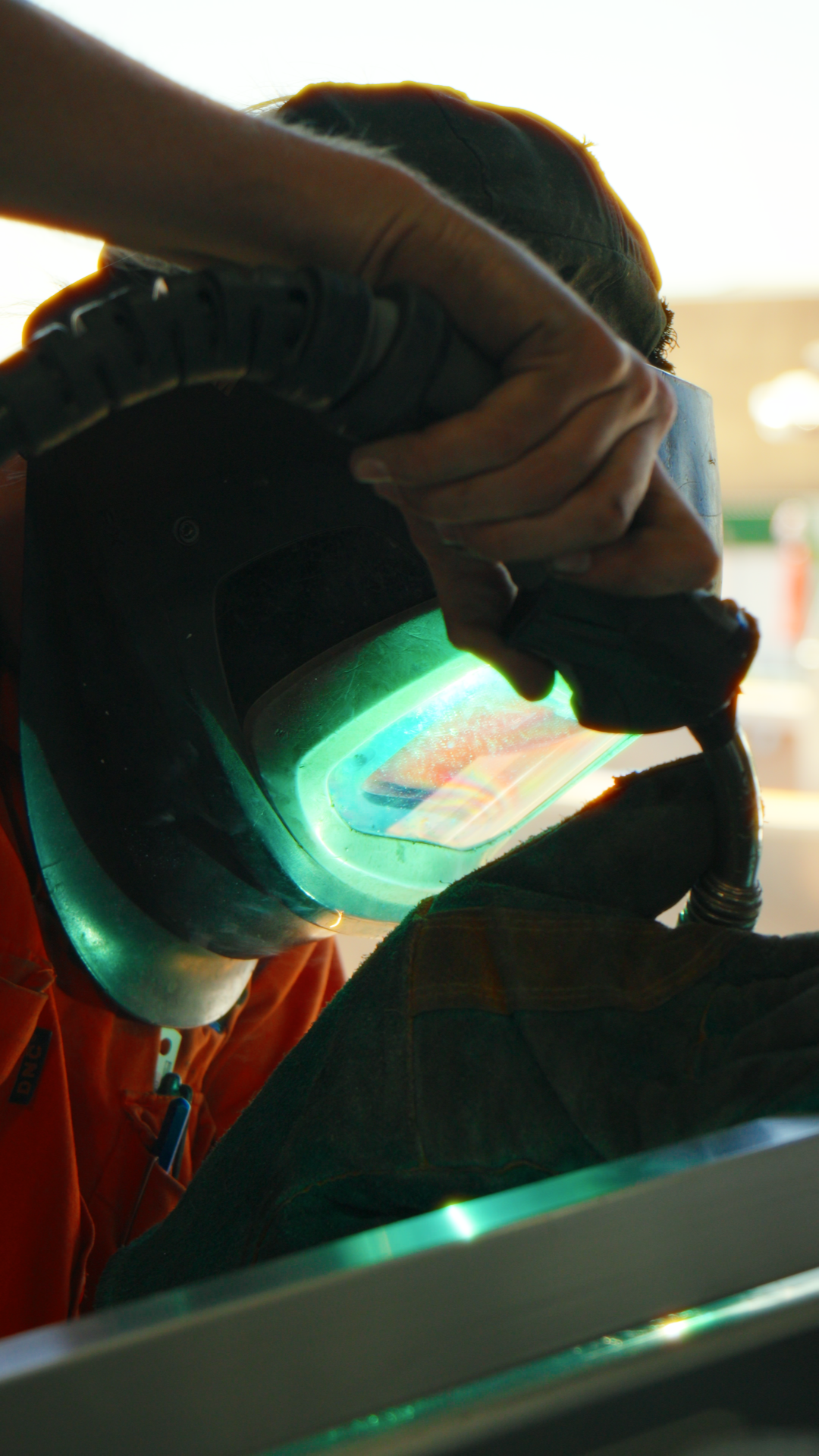 A person wearing safety gloves and an orange jumpsuit is welding, with a bright arc of light visible through a protective helmet with a reflective visor.