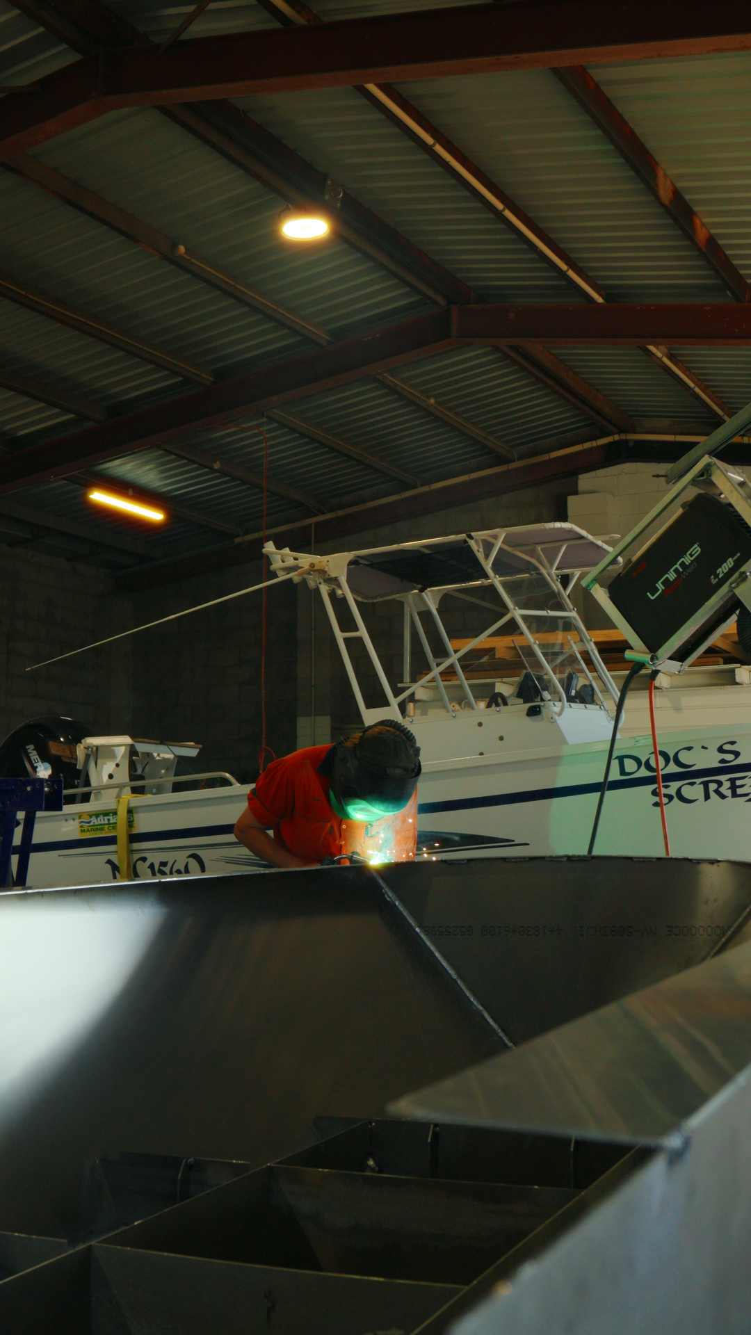 A person welding a metal boat inside a workshop with a boat labeled 'Doc's' and another boat on a lift in the background.