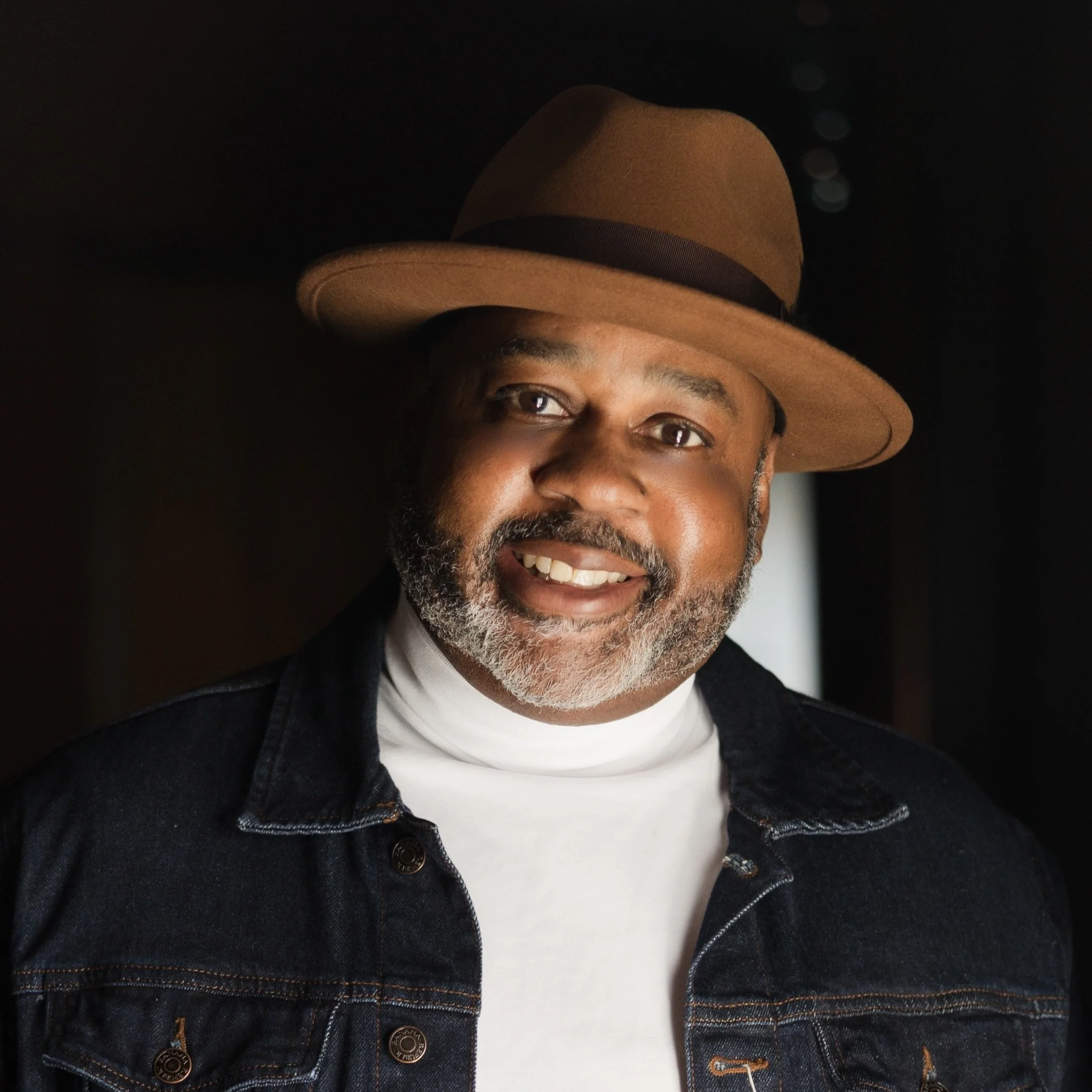 A smiling man wearing a brown fedora hat and a black denim jacket over a white turtleneck, posed indoors with a dark background and soft lighting.