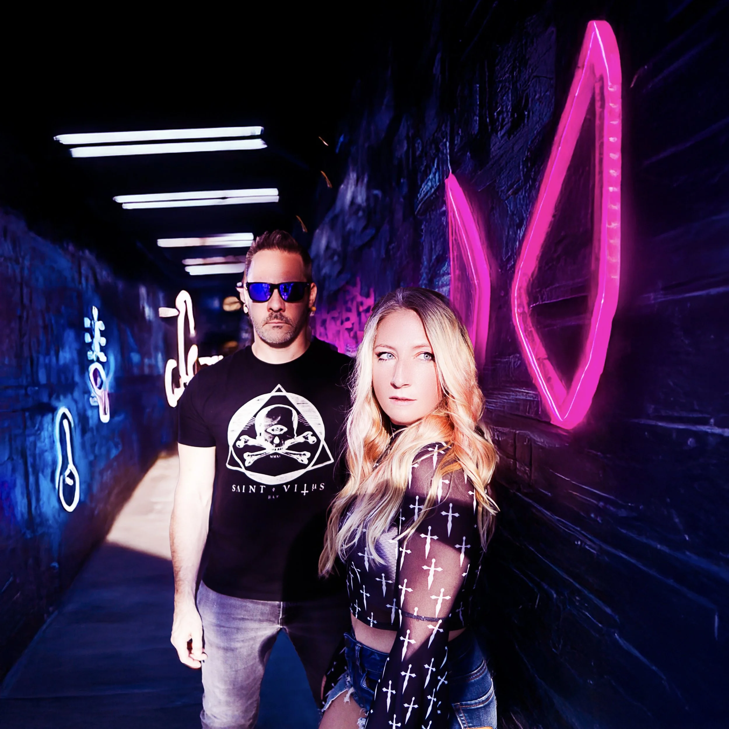 A man with sunglasses and a woman with long blonde hair pose in a neon-lit tunnel decorated with various neon signs, including a large pink geometric shape and several smaller blue and white icons.