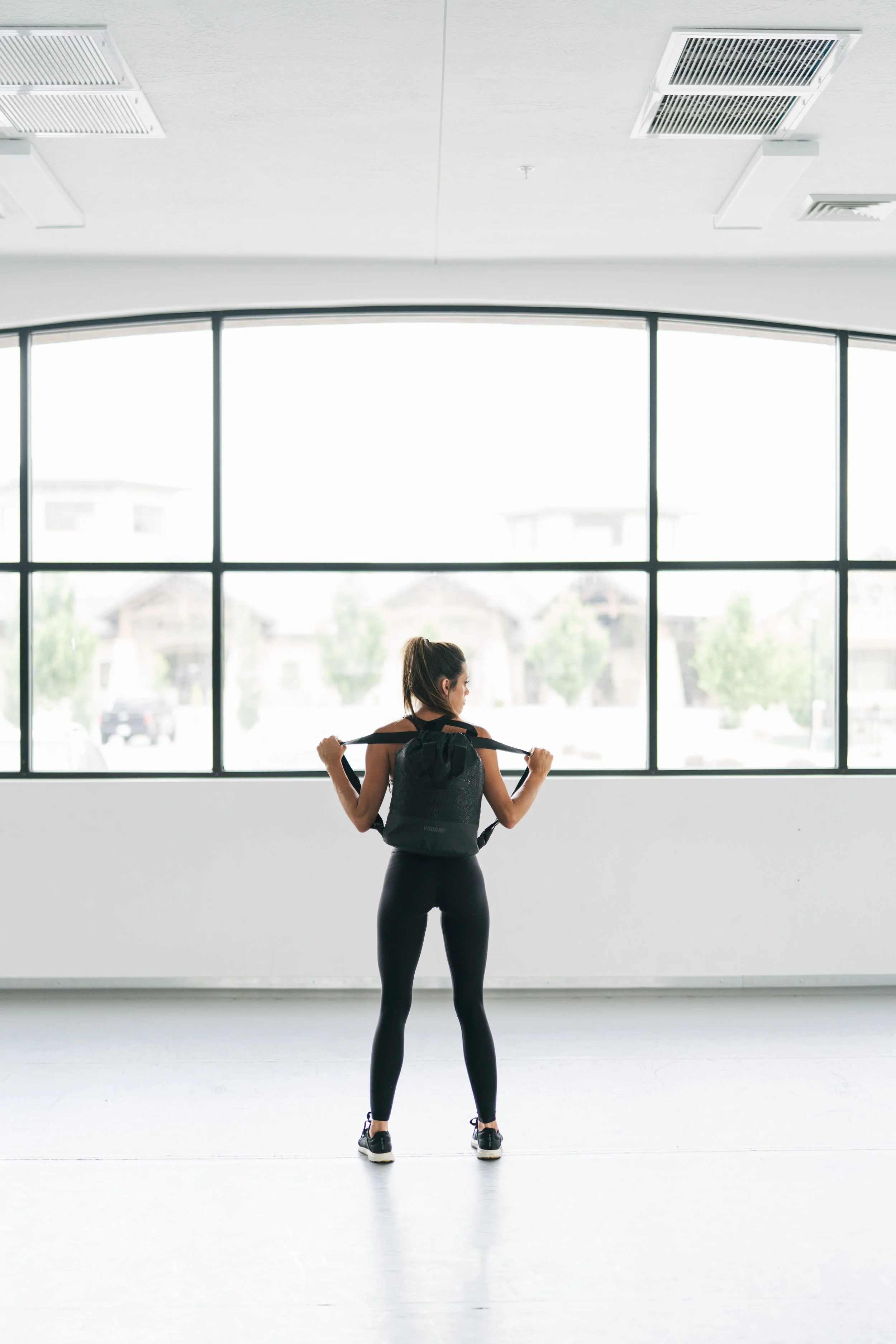 A woman in athletic clothing standing in a bright room with large windows, holding a resistance band across her shoulders, preparing for exercise.