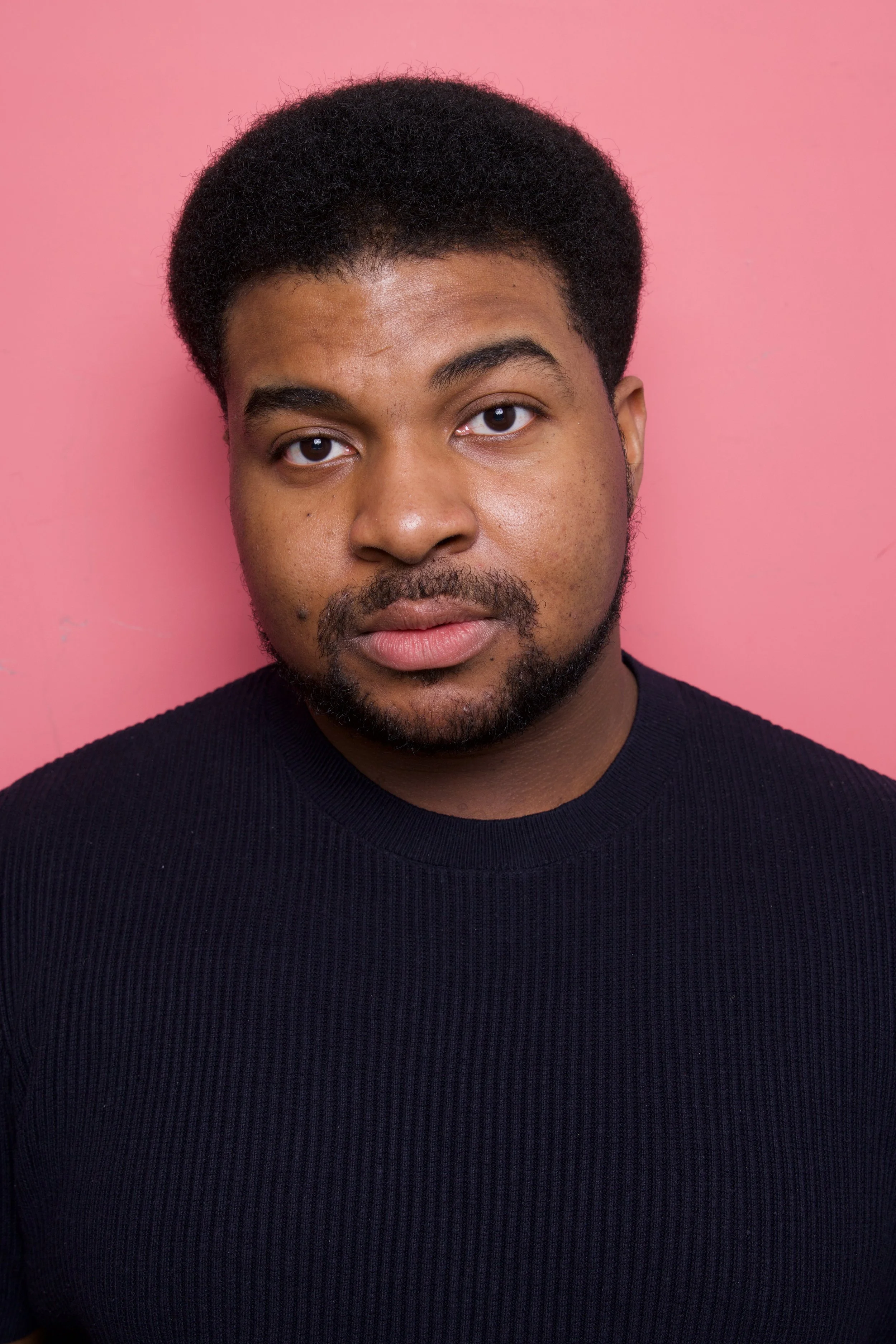Close-up headshot of a young man, Steven Rutledge, with short curly black hair, a beard, and mustache, wearing a black ribbed shirt, standing against a pink background.

Taken by Robert Quiles (@robertredphoto on IG)
