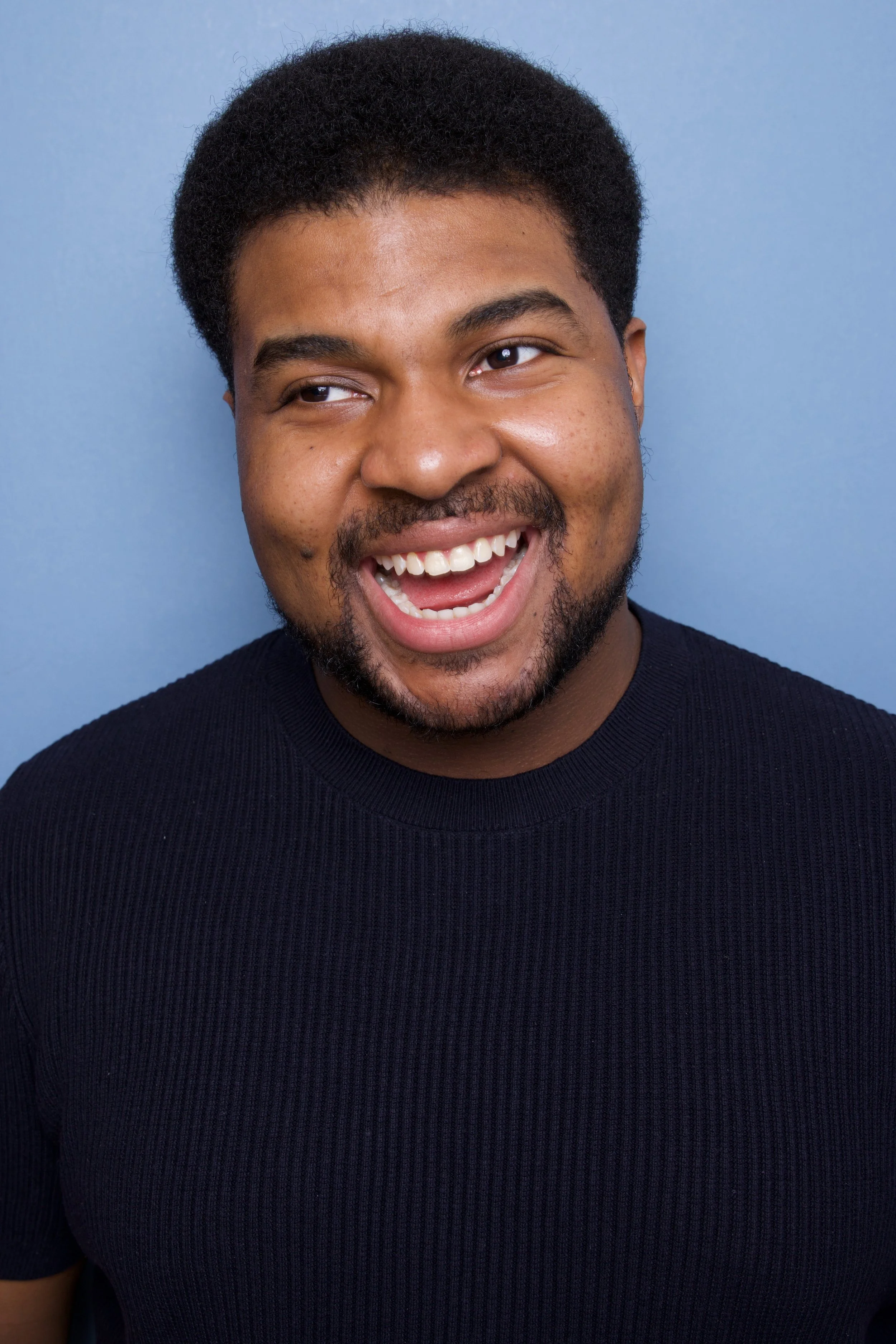 A young man, Steven Rutledge, with short curly hair and a beard, wearing a ribbed black shirt, smiling and looking slightly to the side, against a light blue background.