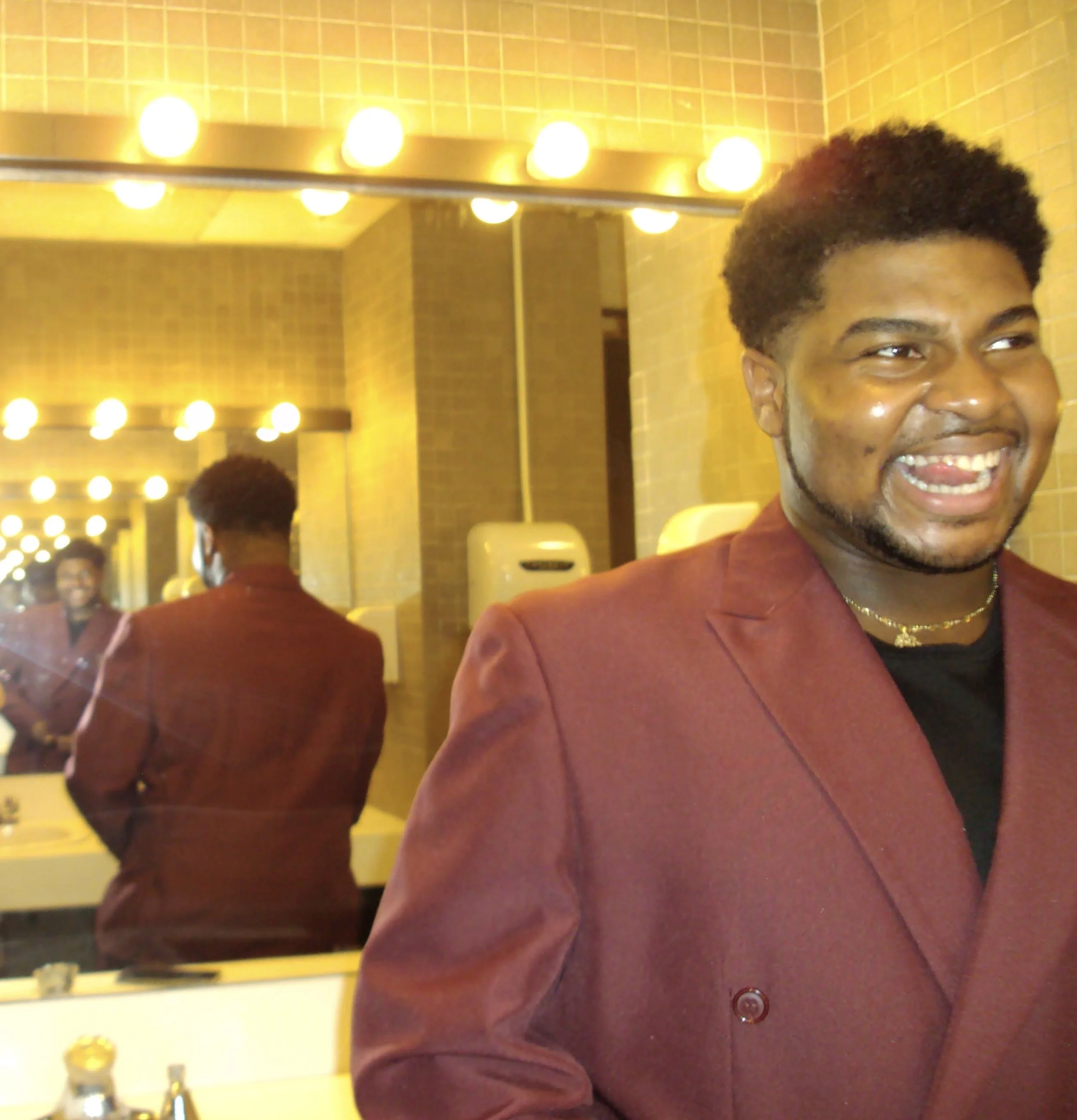 Steven, wearing a burgundy blazer, stands smiling mischievously in front of a mirror