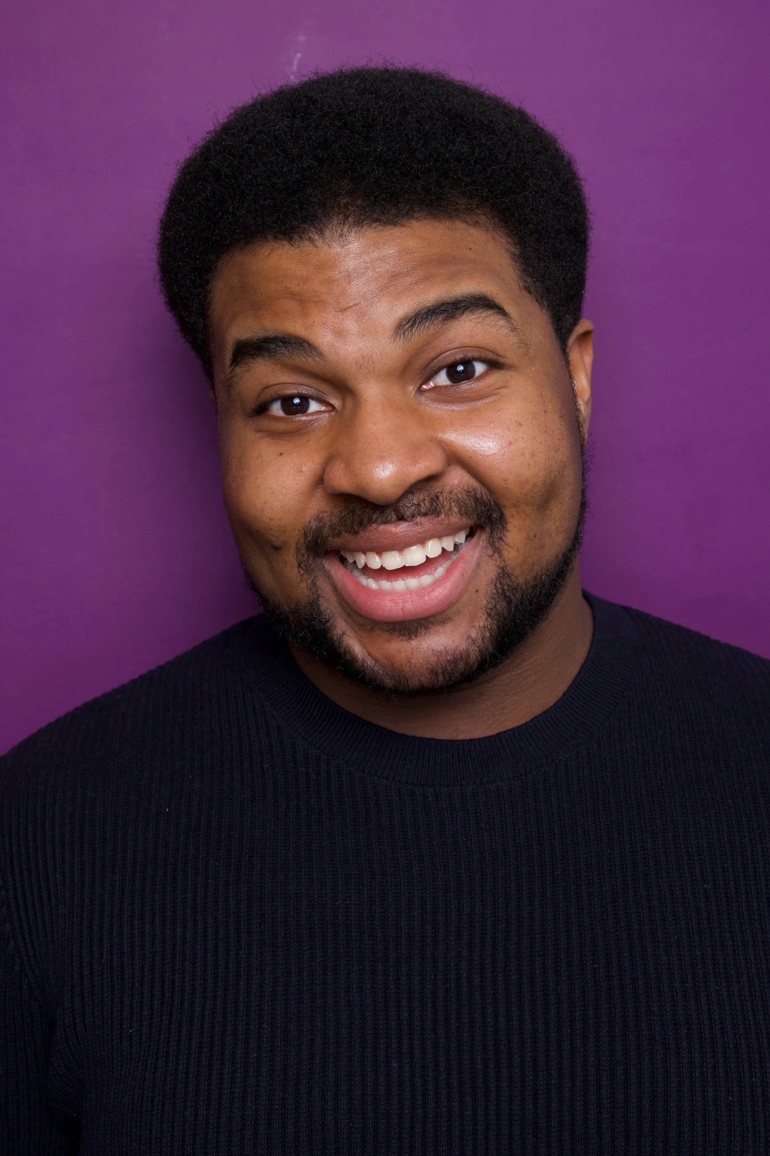 Close-up headshot of a young man, Steven Rutledge, with short curly black hair, a beard, and mustache, wearing a black ribbed shirt, standing against a purple background.

Taken by Robert Quiles (@robertredphoto on IG)