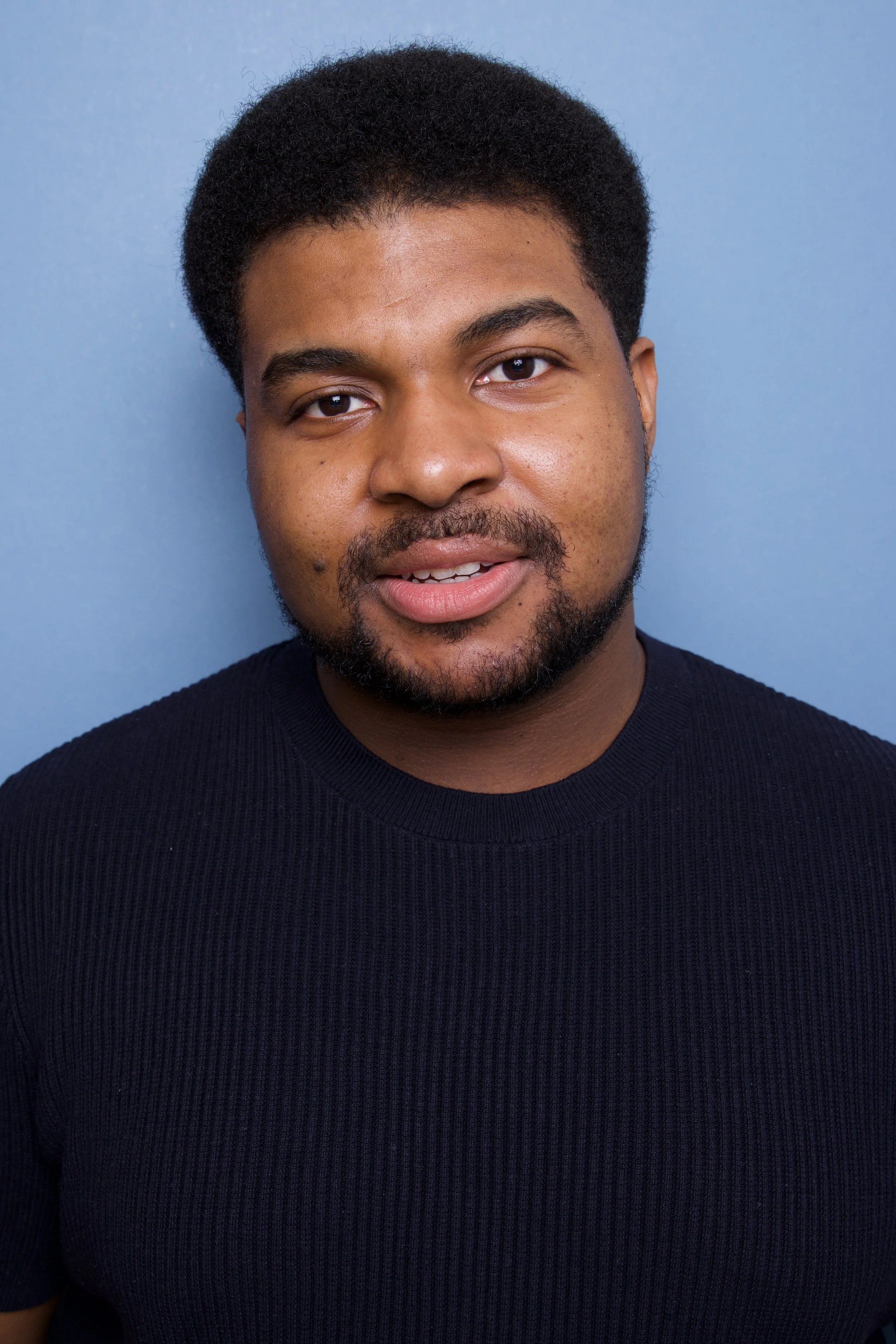 Close-up headshot of a young man, Steven Rutledge, with short curly black hair, a beard, and mustache, wearing a black ribbed shirt, standing against a light blue background.

Taken by Robert Quiles (@robertredphoto on IG)