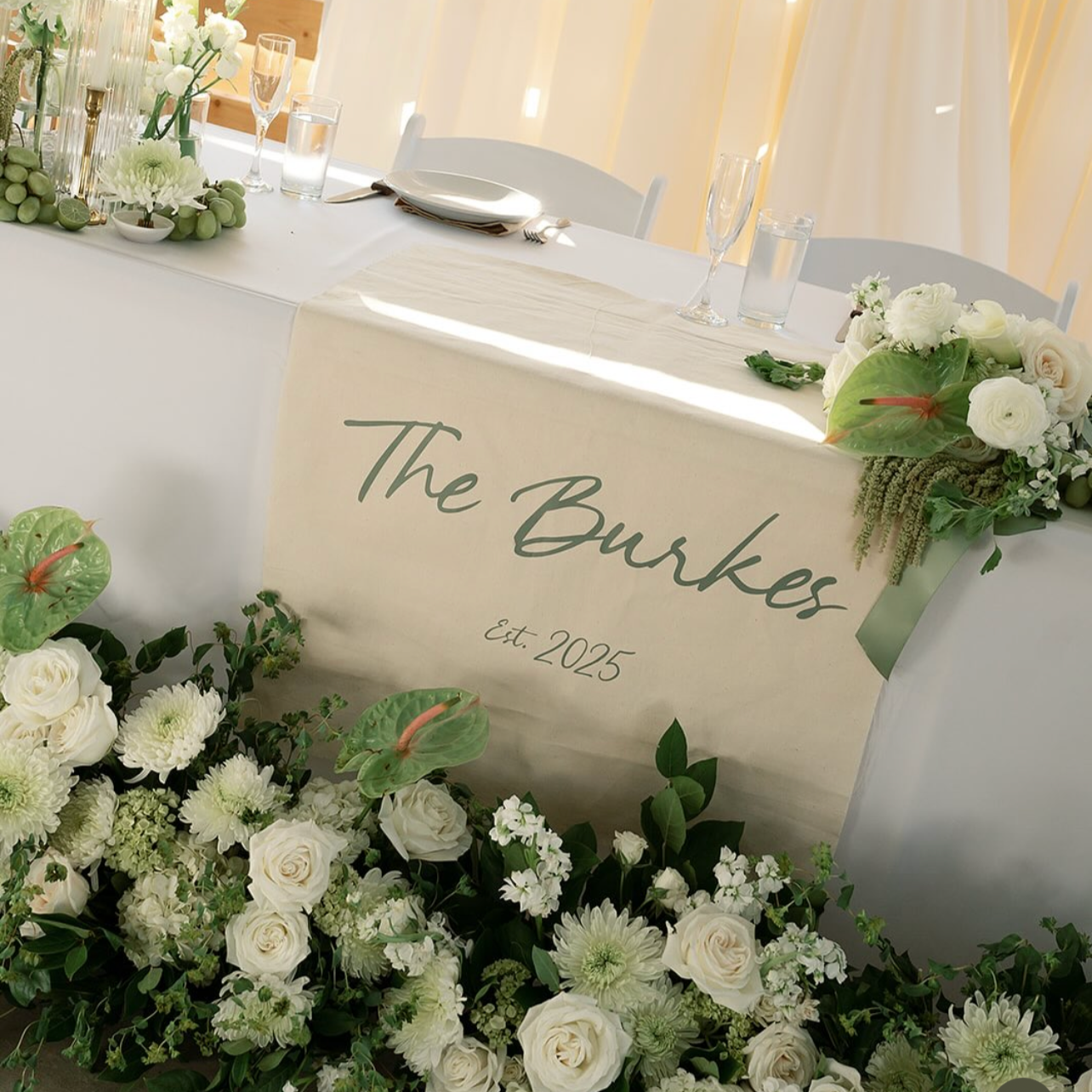 Decorated outdoor wedding reception table with white flowers, greenery, and a sign reading "The Burkes Est. 2025", set against a wooden privacy fence.