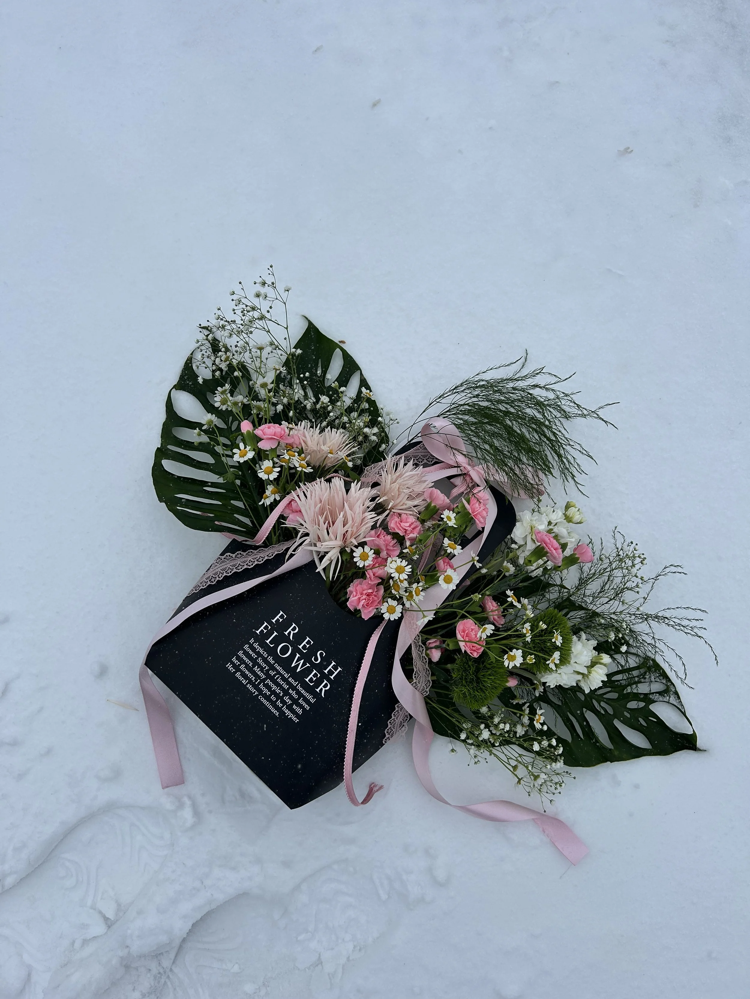 Pink and white flowers in a black box with 'Fresh Flower' written on it, surrounded by large green leaves and white snow.
