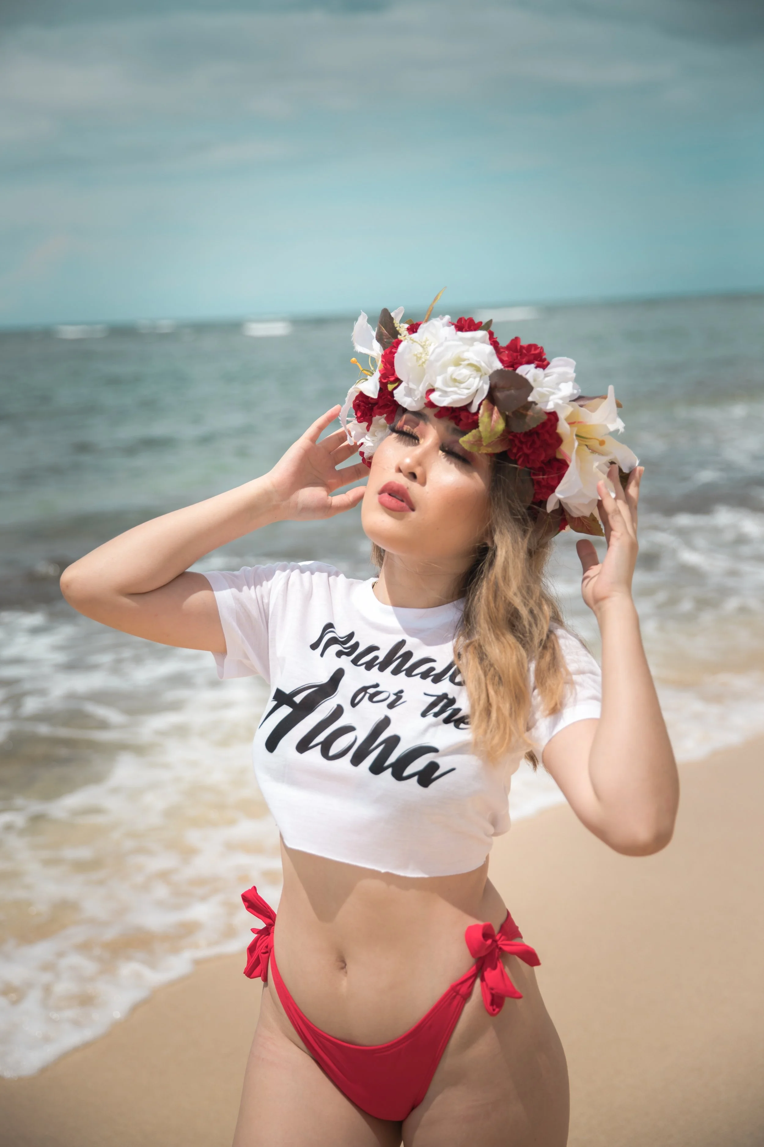 A woman on the beach wearing a floral crown, a white crop top that says 'Mahalo for the Aloha,' and red bikini bottoms with bows, standing with her eyes closed and hand near her temple.