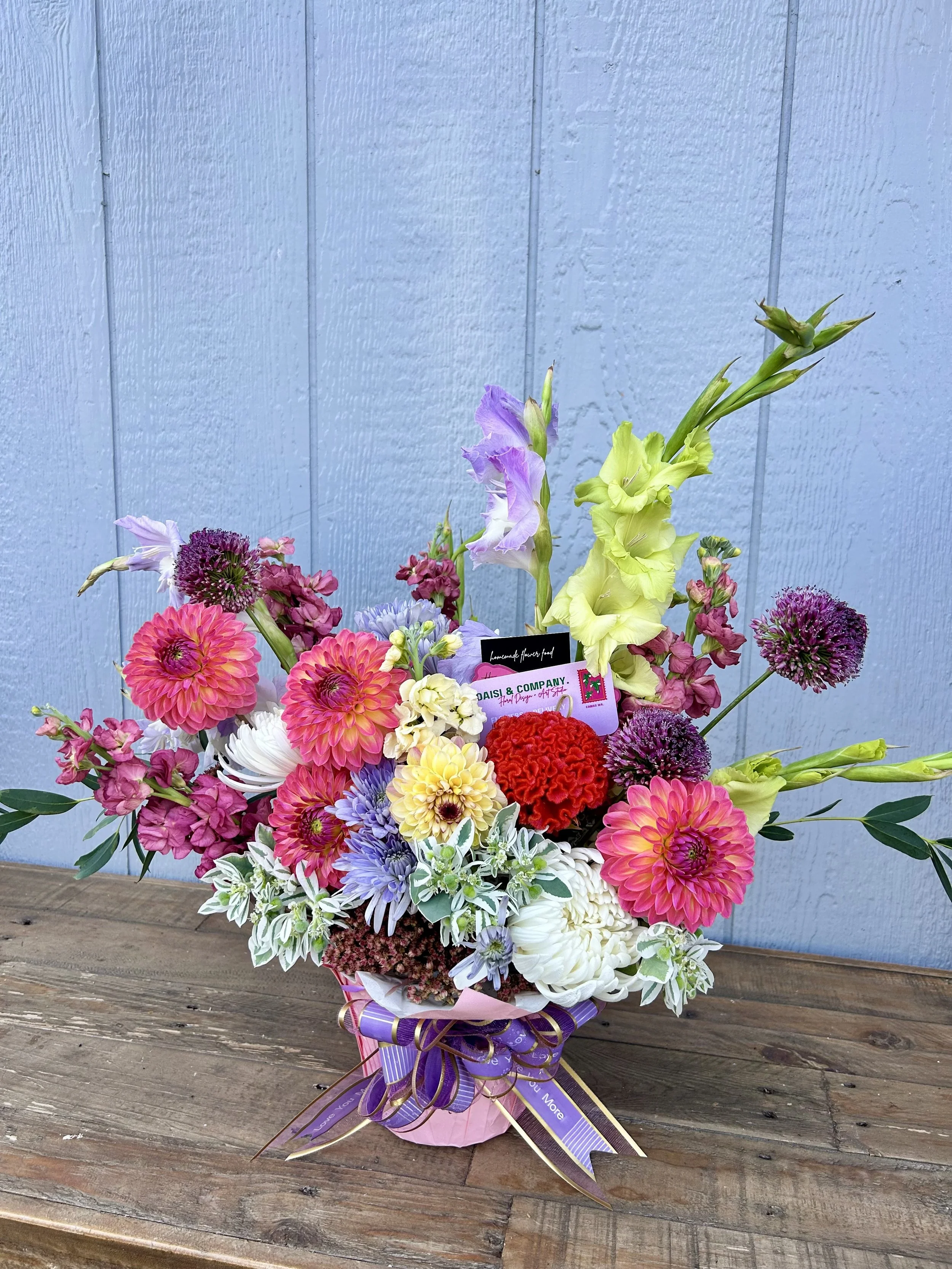 Colorful mixed flower bouquet with pink, purple, white, yellow, and red flowers in a pink wrapper with purple ribbons, placed on a wooden surface against a light blue wall.