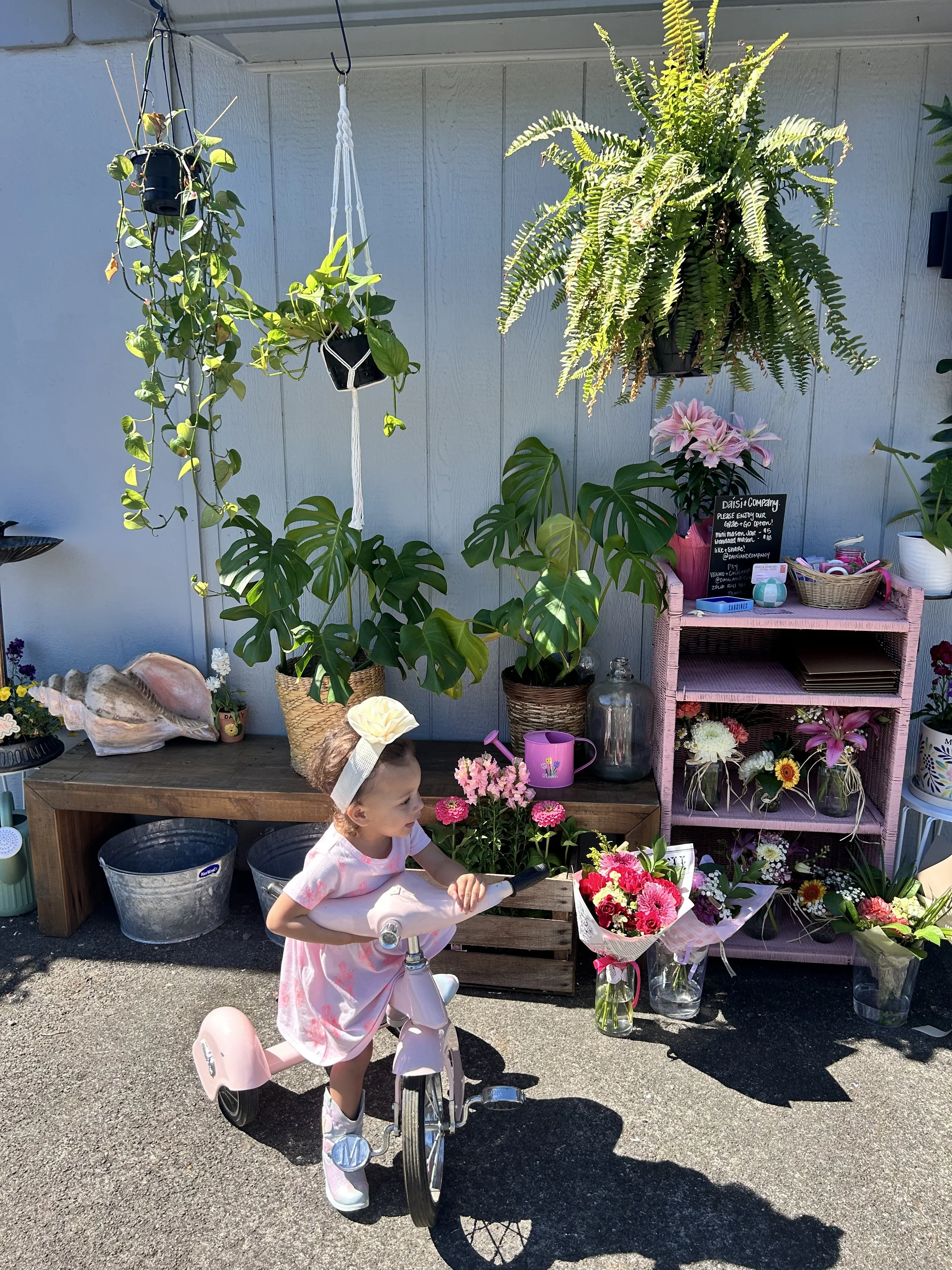 A young girl riding a pink scooter outdoors near a flower stand with various colorful flowers, plants, and decorative items against a light gray wall.