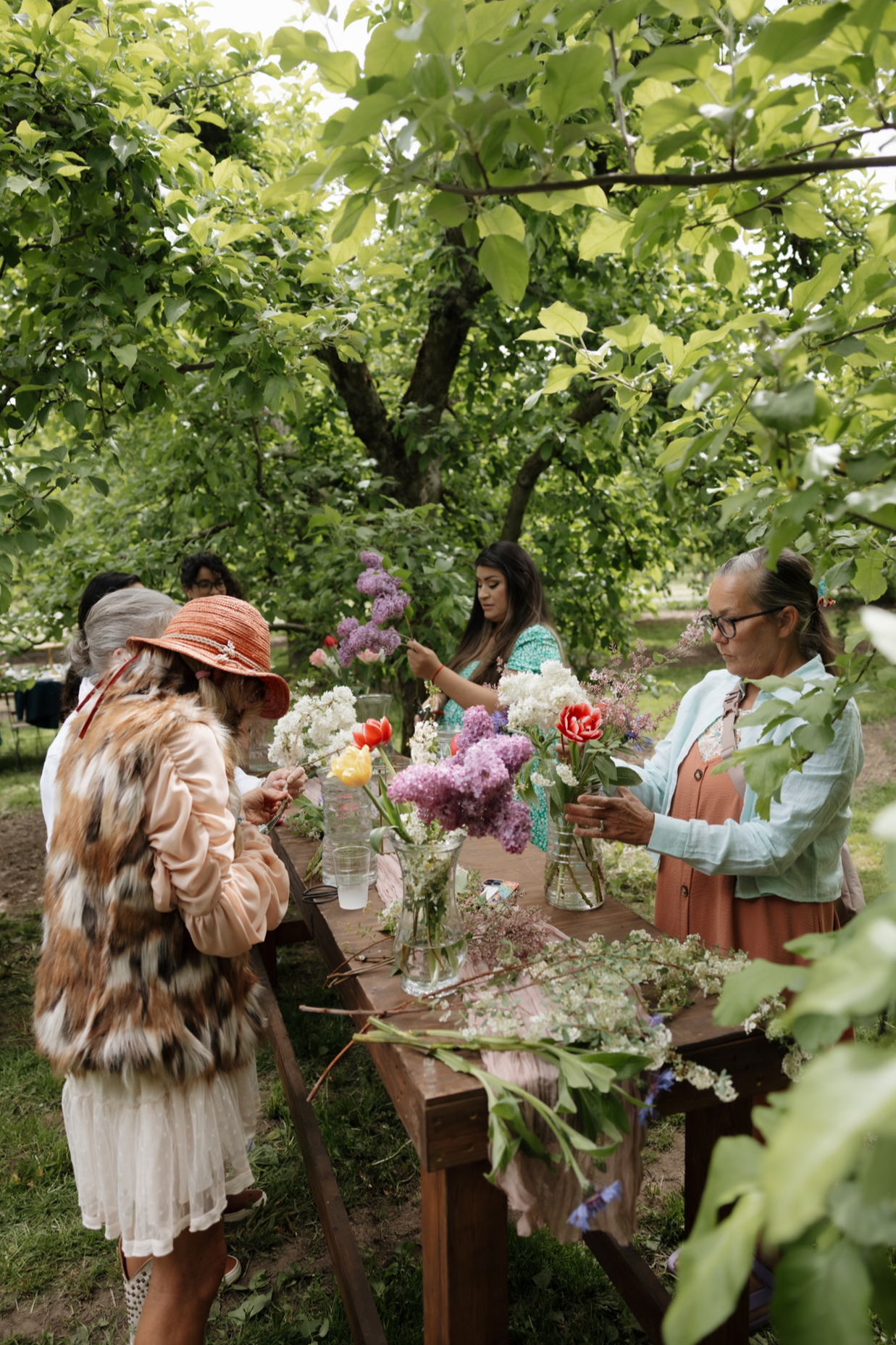 A group of women arranging colorful flowers in vases during a flower arranging workshop outdoors beneath leafy trees.