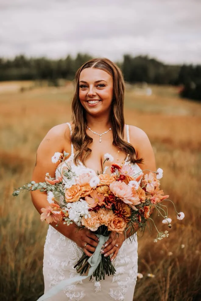 A woman in a white lace wedding dress holding a large bouquet of peach, pink, and white flowers with greenery outdoors in a field on a cloudy day.