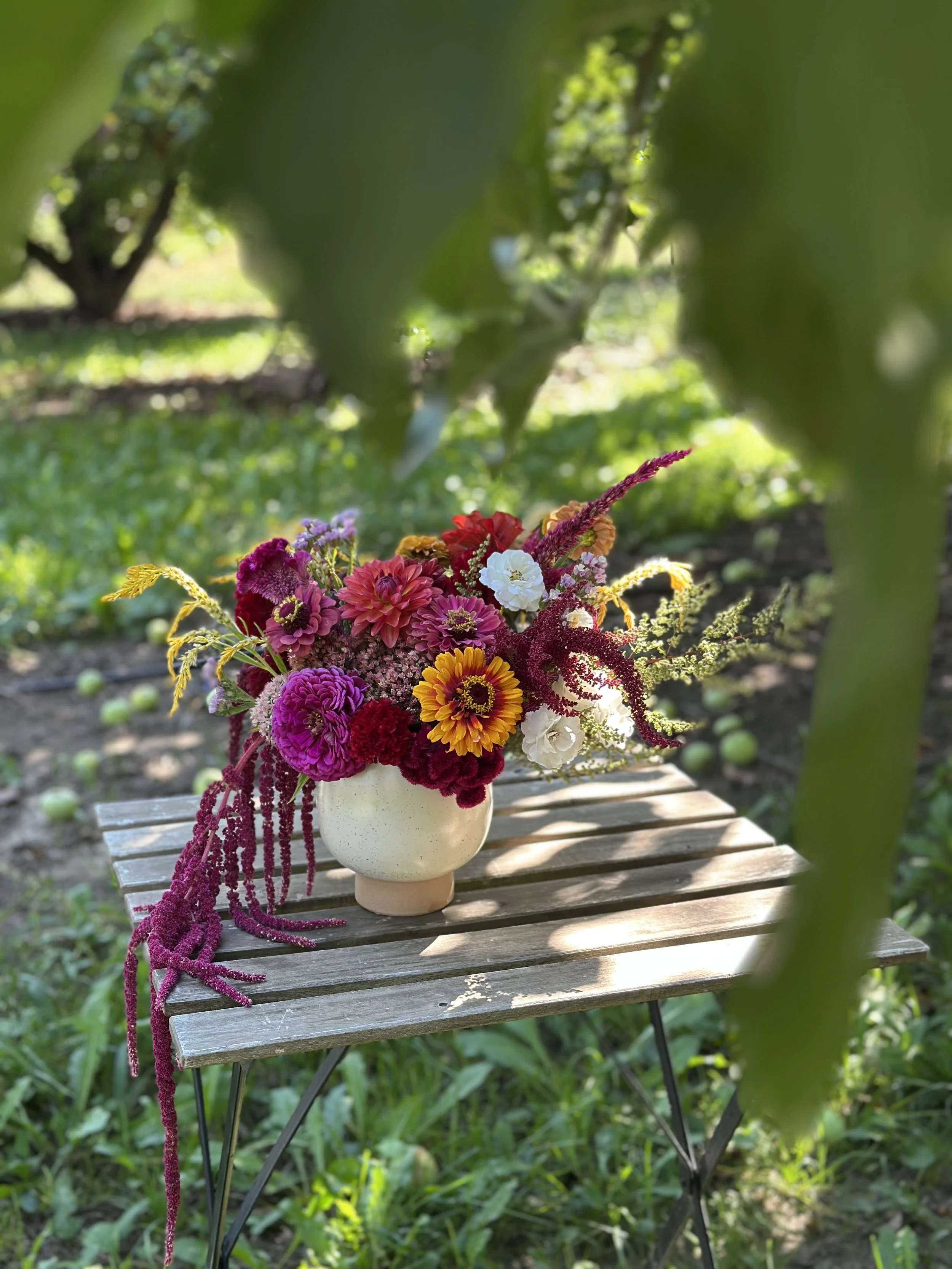 A bouquet of colorful flowers in a white vase placed on a small wooden table outdoors with green foliage and trees in the background.