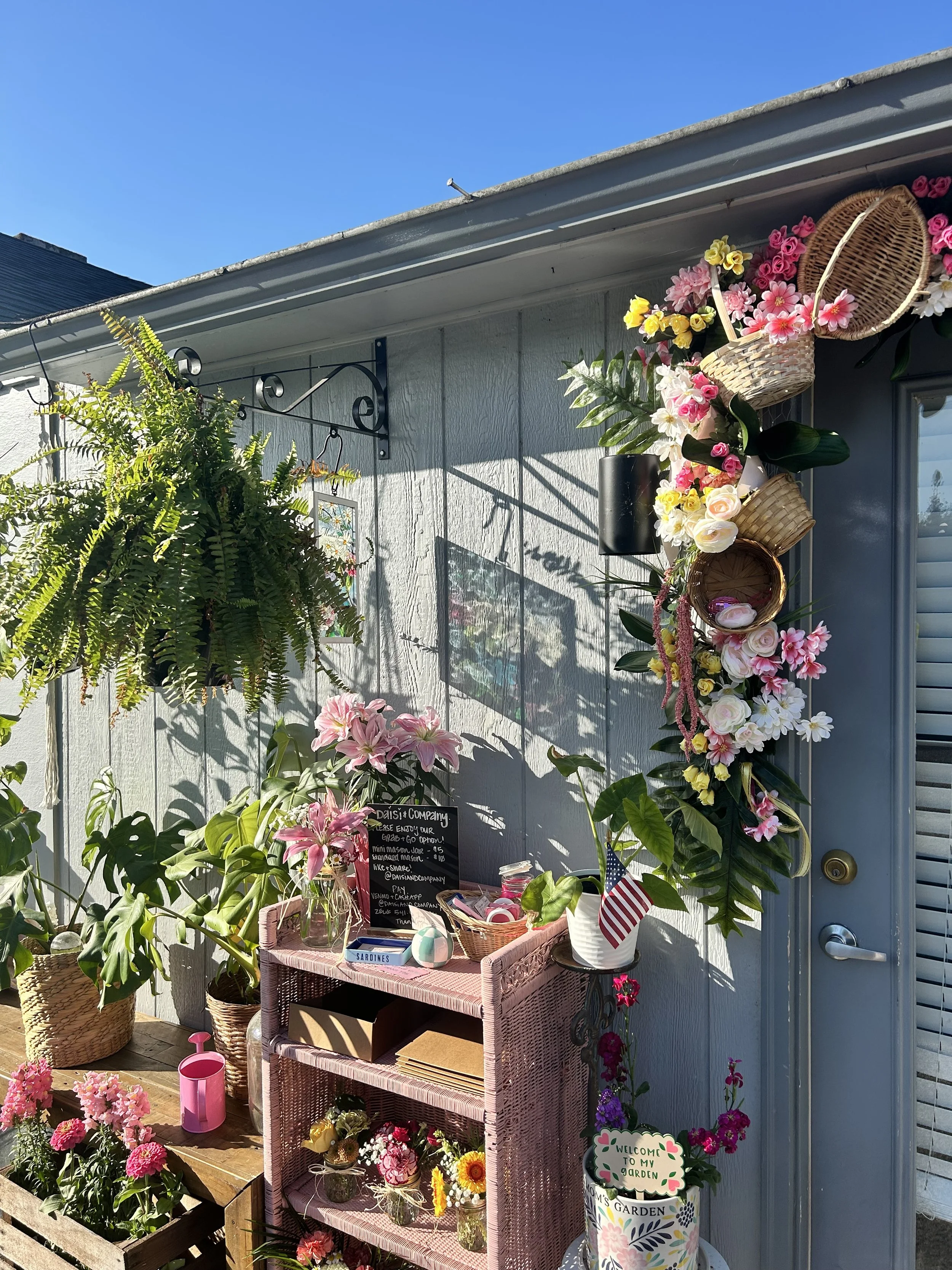 Front porch decorated with colorful flowers, baskets, potted plants, and a pink shelf holding small flower arrangements. Bright sun and clear blue sky overhead.