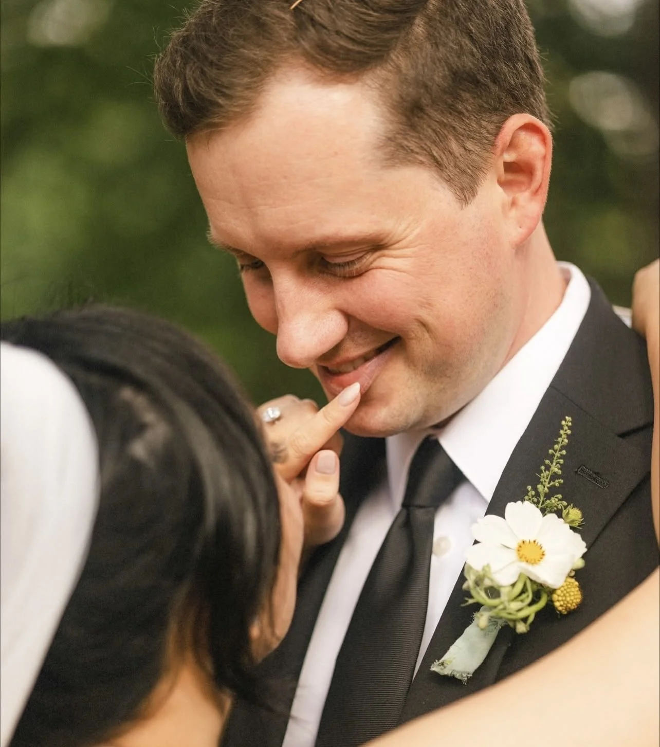 A man in a tuxedo with a flower boutonniere smiles as a woman touches his lips with her finger during what appears to be a wedding or romantic moment.