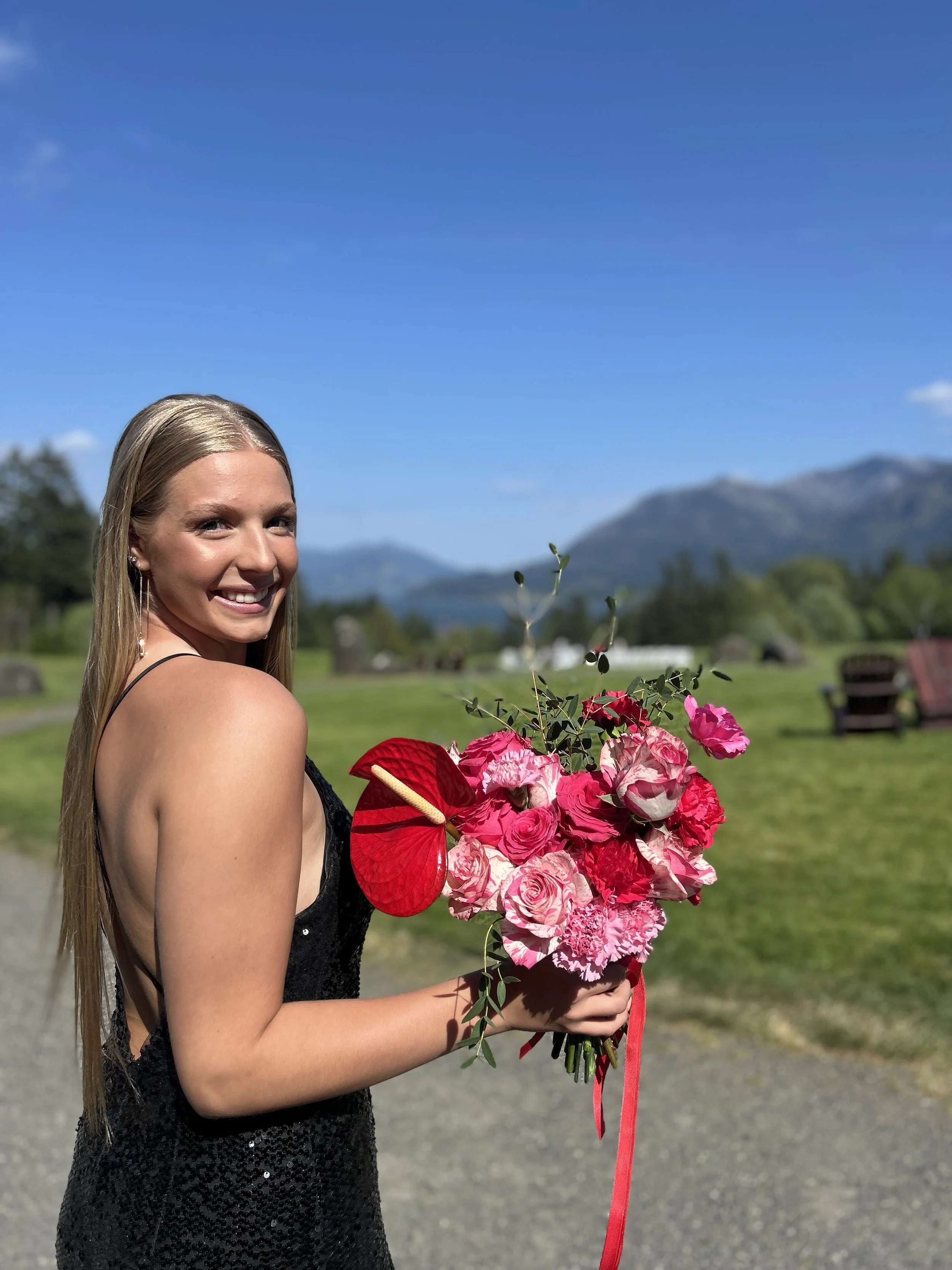 A young woman in a black dress holding a bouquet of pink and red flowers, standing outdoors on a sunny day with mountains in the background.