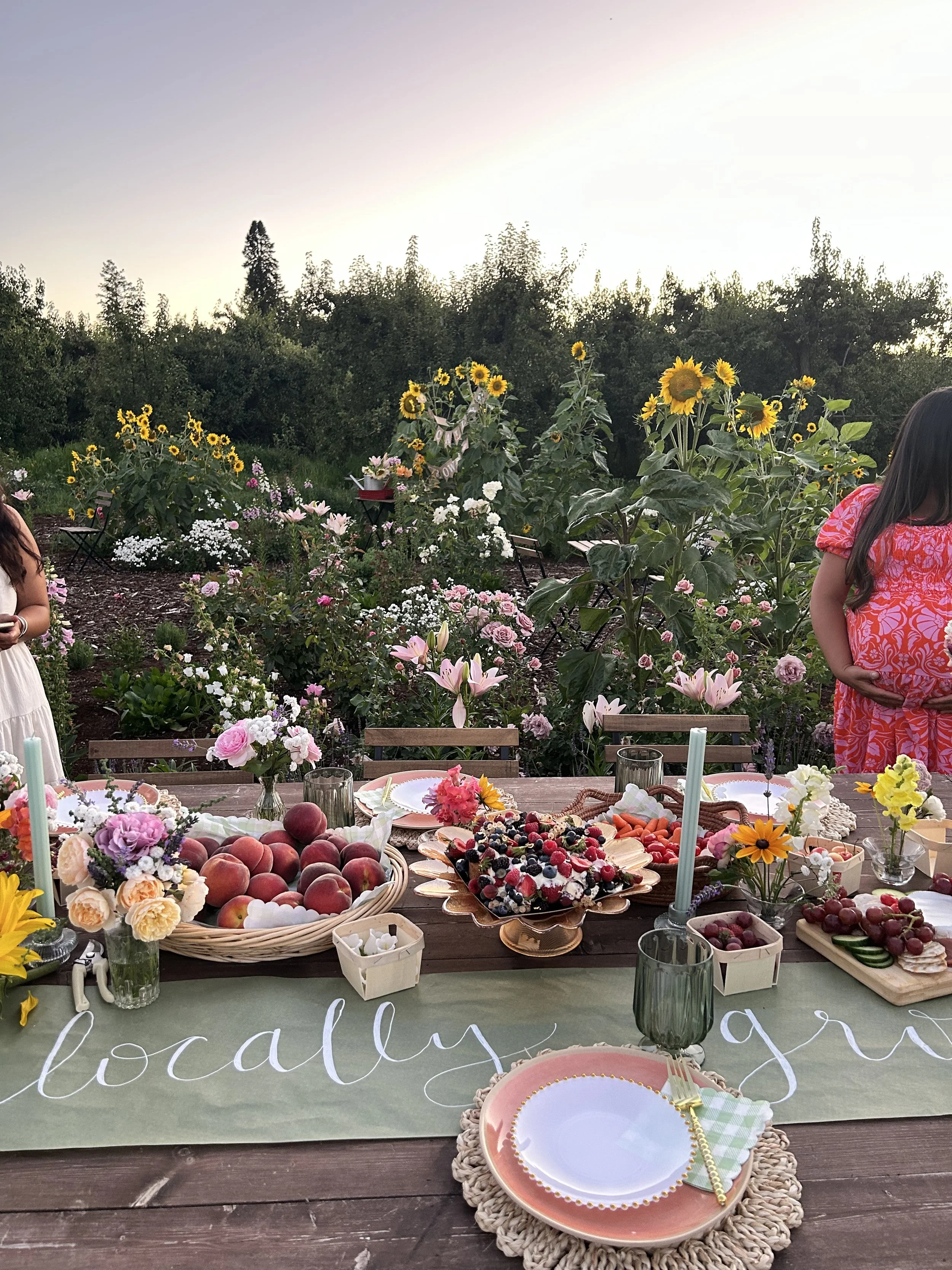 An outdoor garden reception table with fruits, berries, flowers, and tableware, set amidst blooming flowers and sunflowers at sunset.